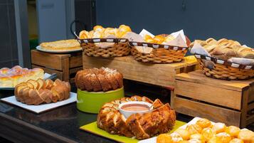 A buffet table with various breads and pastries, including sliced bread, rolls, and a loaf of bread in a green container.