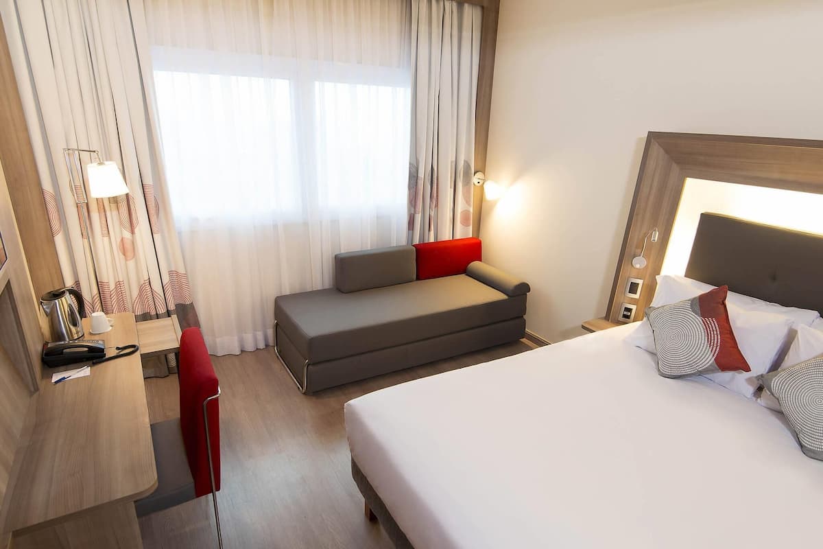 A hotel room with a bed, a desk with a chair, a sofa, and a window with curtains.
