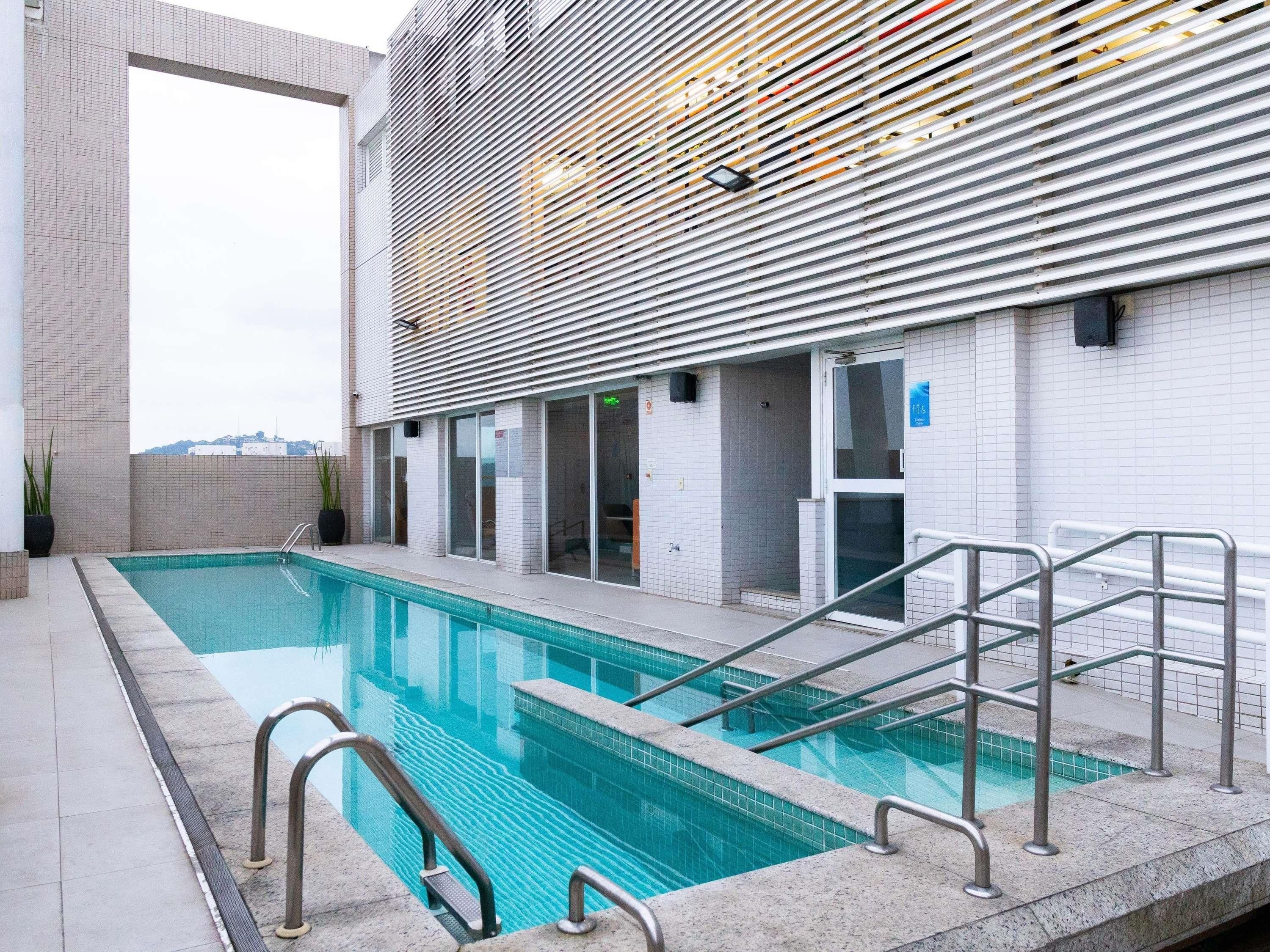 Une piscine avec une terrasse carrelée, des rampes métalliques et, au fond, un bâtiment dont la façade est recouverte de lambris.