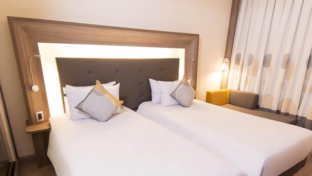 A hotel room with two beds, a wooden headboard, a nightstand, a lamp, and a window with curtains. - Novotel Santos Gonzaga (Santos)