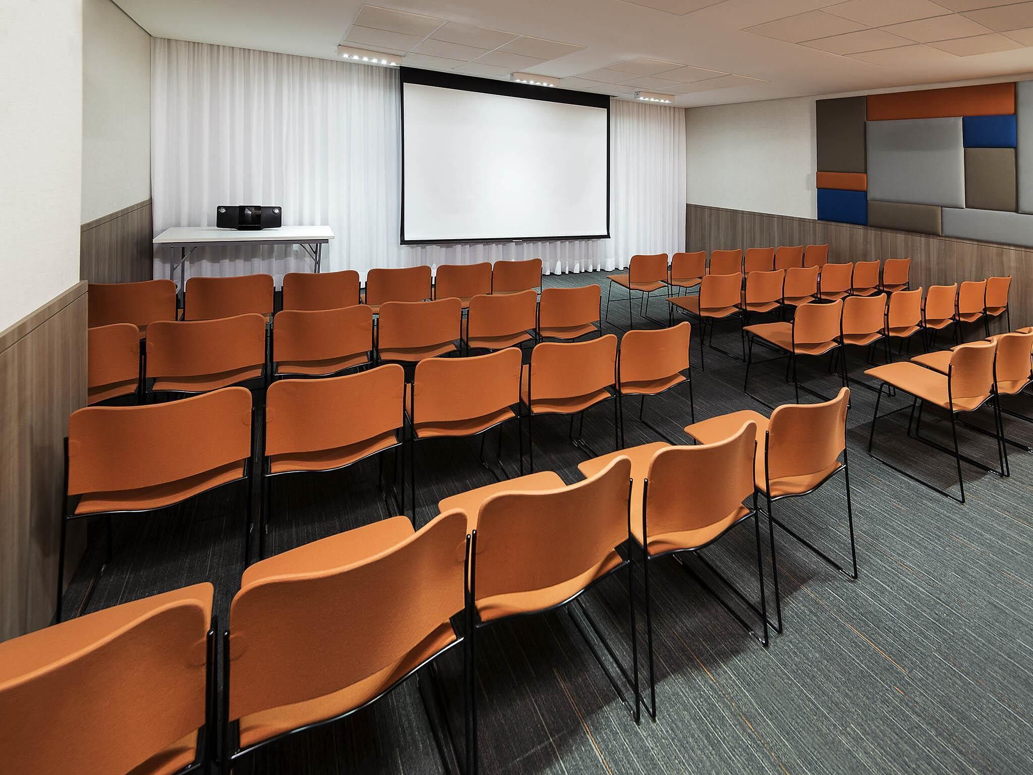 A conference room with rows of orange chairs, a projector screen, and a whiteboard.