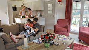 Ein Wohnzimmer mit einer Couch, einem Couchtisch mit Zeitschriften und einer Kerze, einem roten Sessel und einer Küchenzeile, in der jemand kocht.