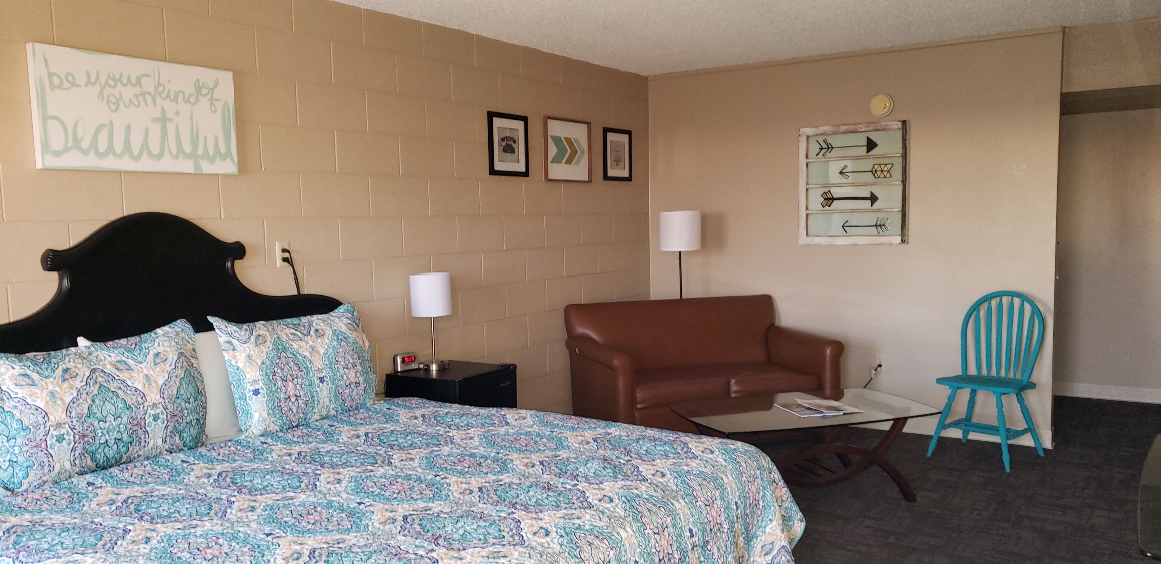 A bedroom with a bed, a brown sofa, a blue chair, a small table, and a wall with framed pictures and a sign.
