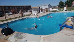 A swimming pool with people swimming and playing, surrounded by a fence, with buildings and a parking area in the background.