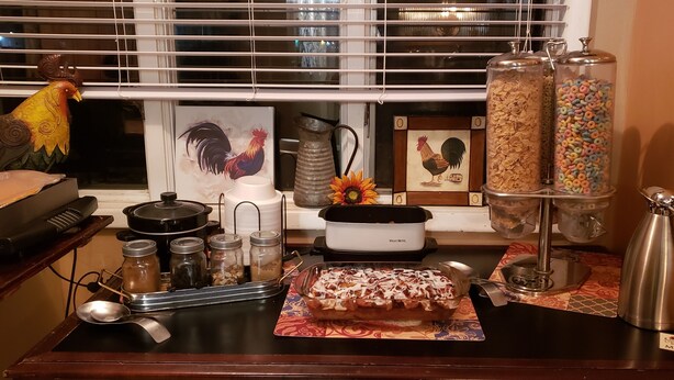 A kitchen counter with a West Bran bread maker, jars of condiments, a coffee pot, and a tray of food. - Kansas Country Inn (Oakley)