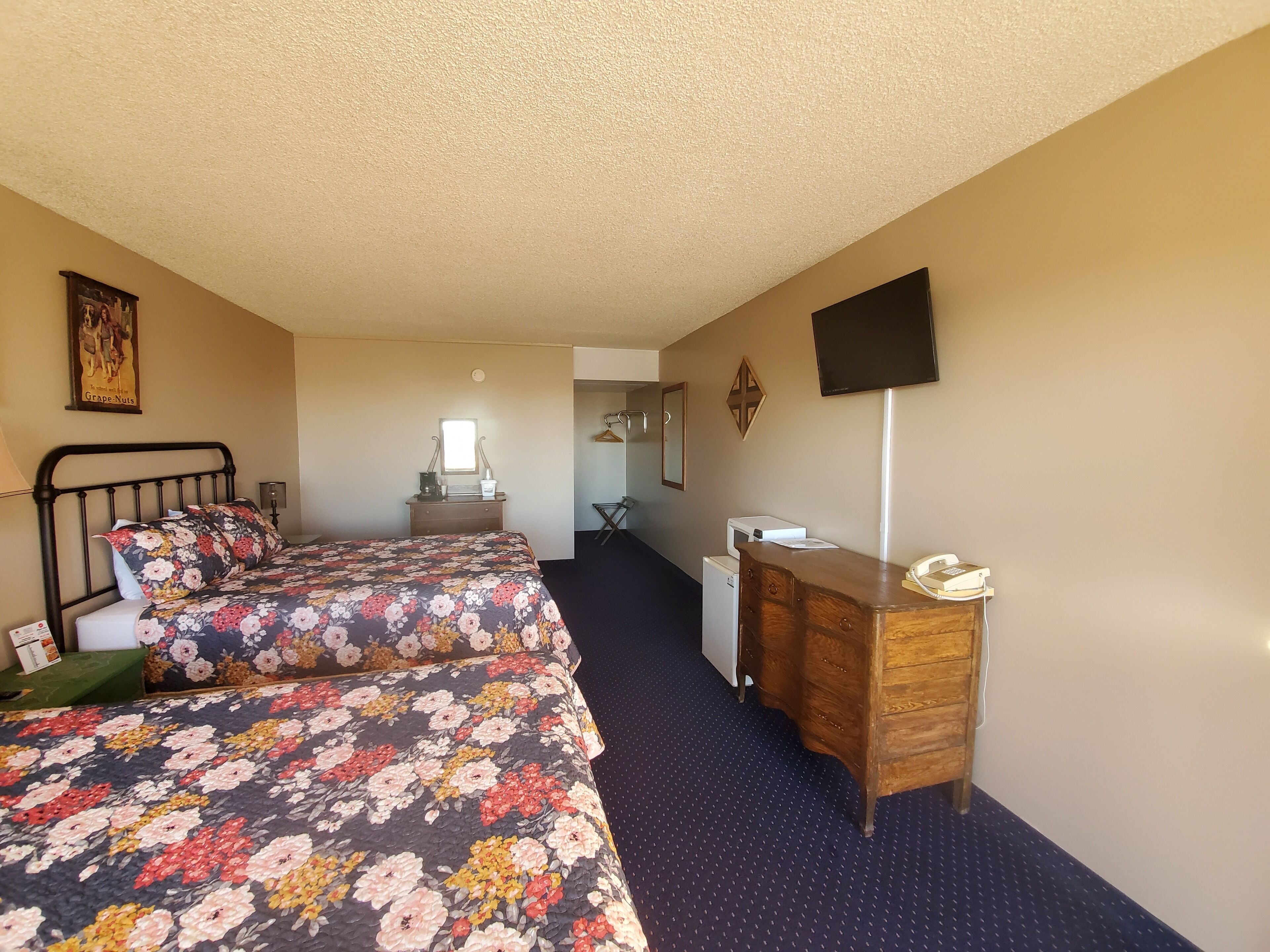 Habitación de hotel con dos camas, un escritorio, un espejo, un televisor y una pequeña cocina.