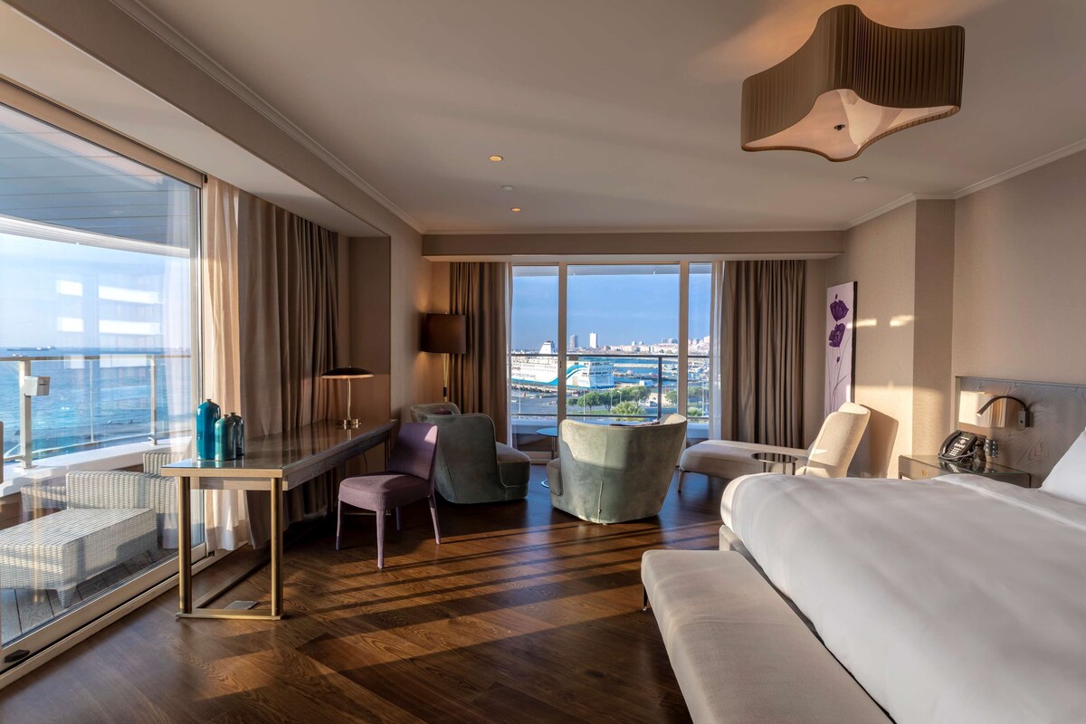 A hotel room with a large bed, a desk, a chair, a sofa, and a view of the city.