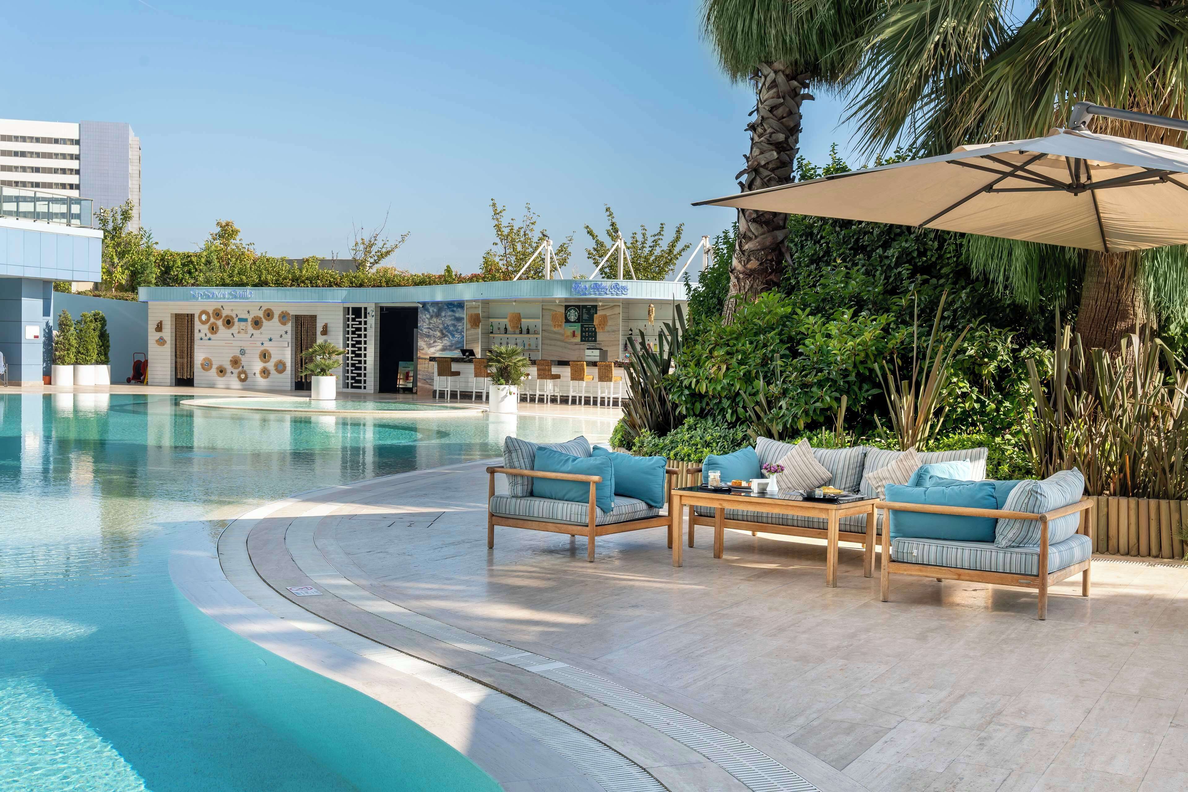 A poolside area with a lounge chair and a table, surrounded by greenery and a building with a sign.