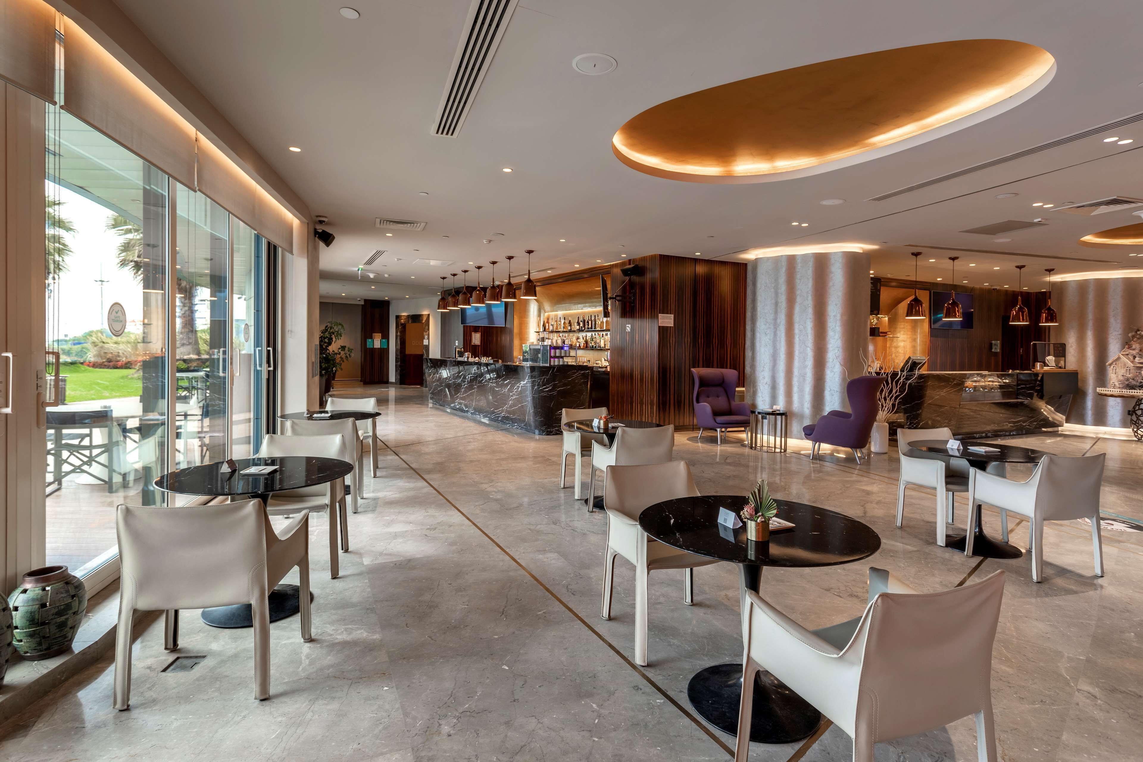 A modern hotel lobby with round tables and chairs, a bar area, and a large window offering a view of the outdoors.