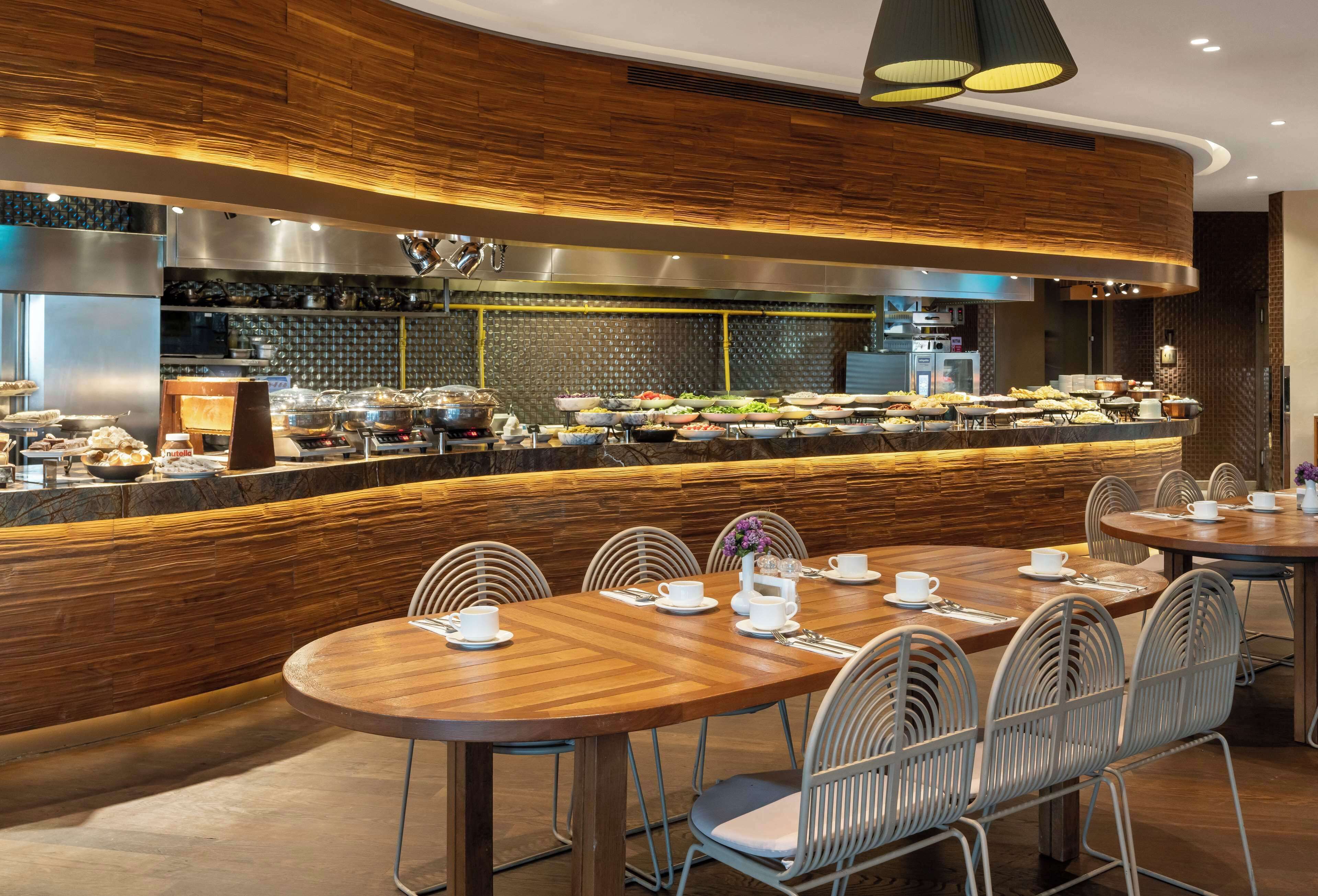 A modern dining area with wooden tables and chairs, a buffet with various food items, and a kitchen in the background.