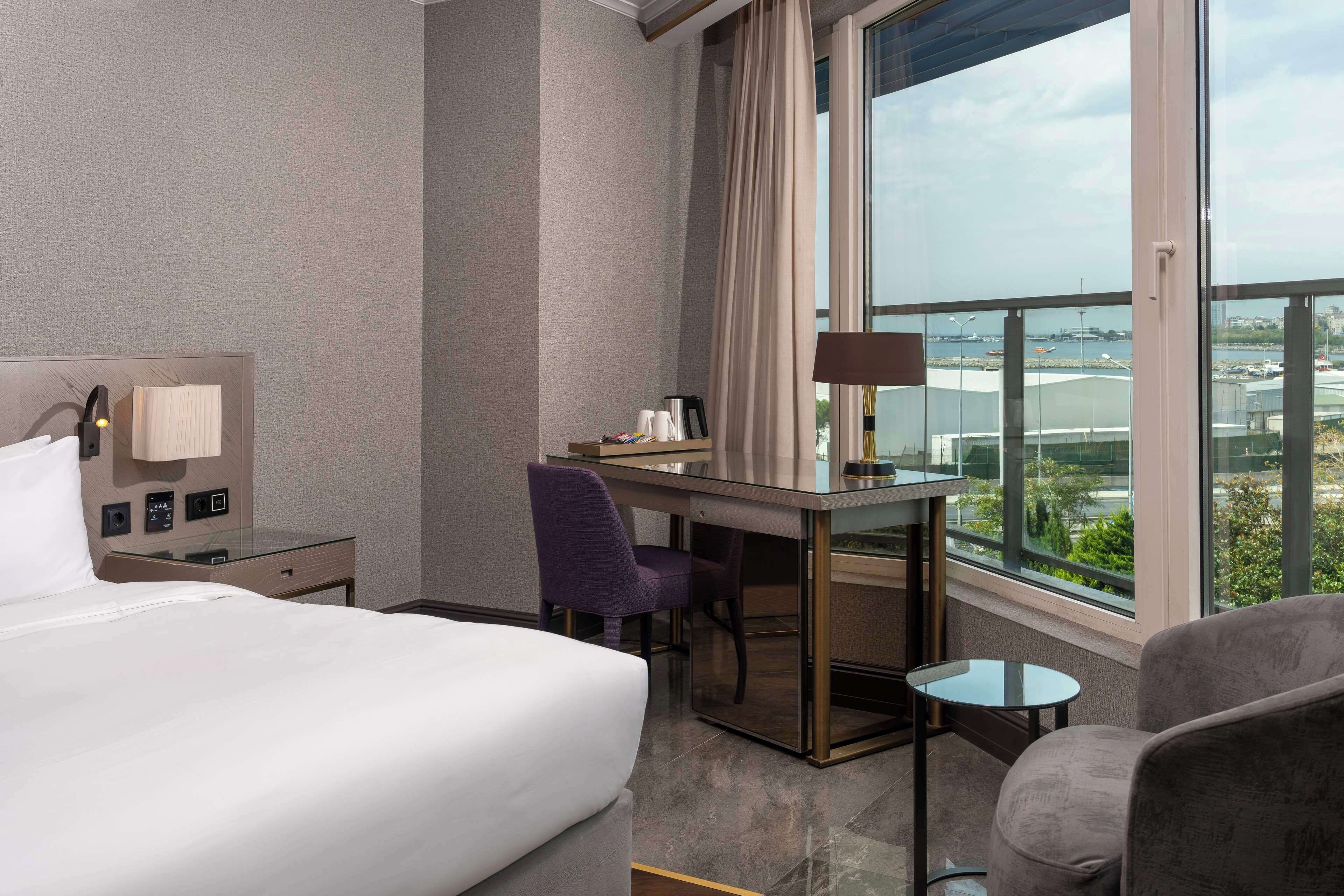 A hotel room with a bed, a desk, a chair, a small table, and a view of the city through large windows.