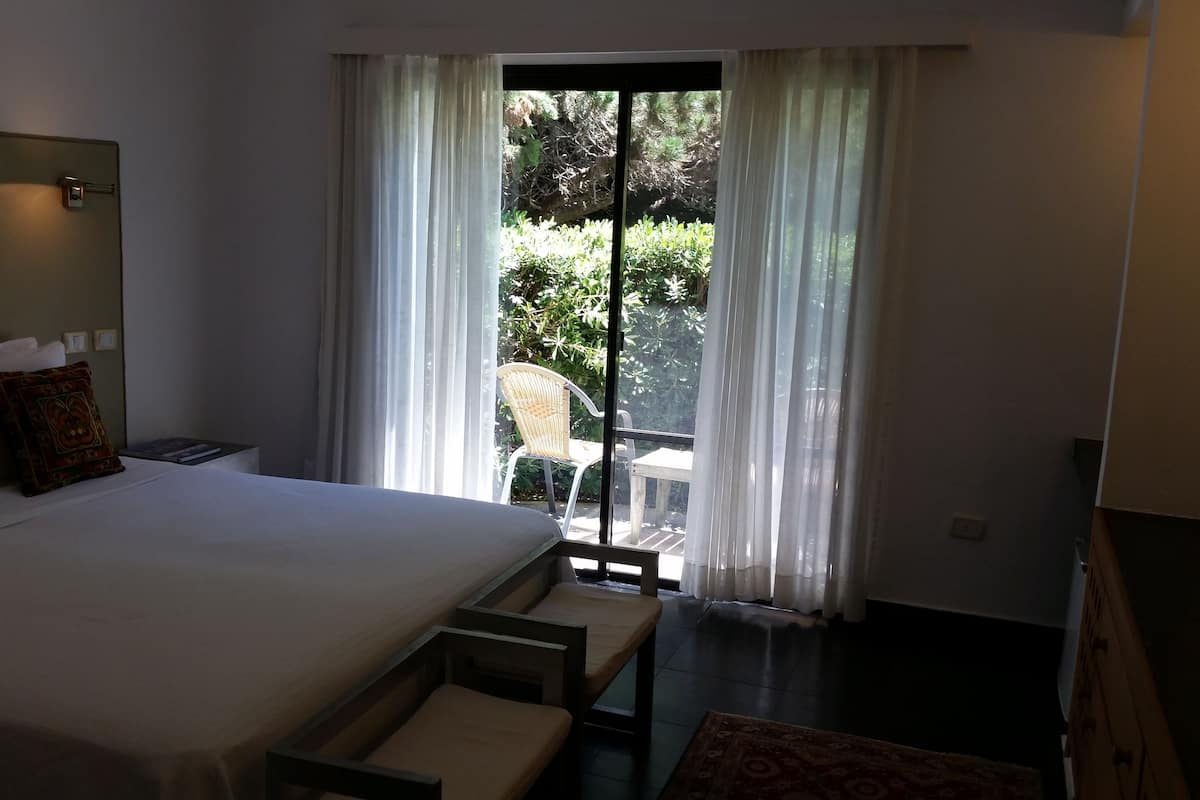 A hotel room with a large bed, a nightstand, a chair, and a view of greenery through the window.