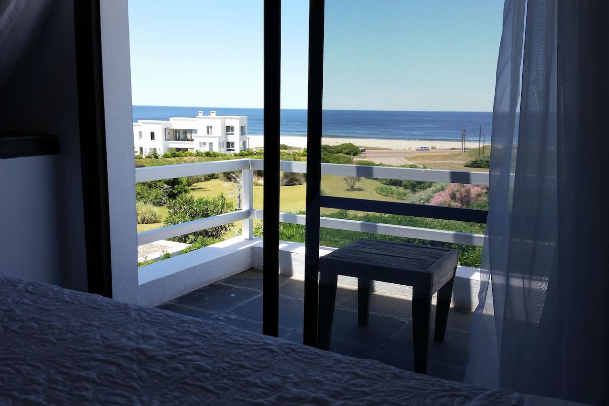 A balcony with a view of the sea, a white building, and a chair.