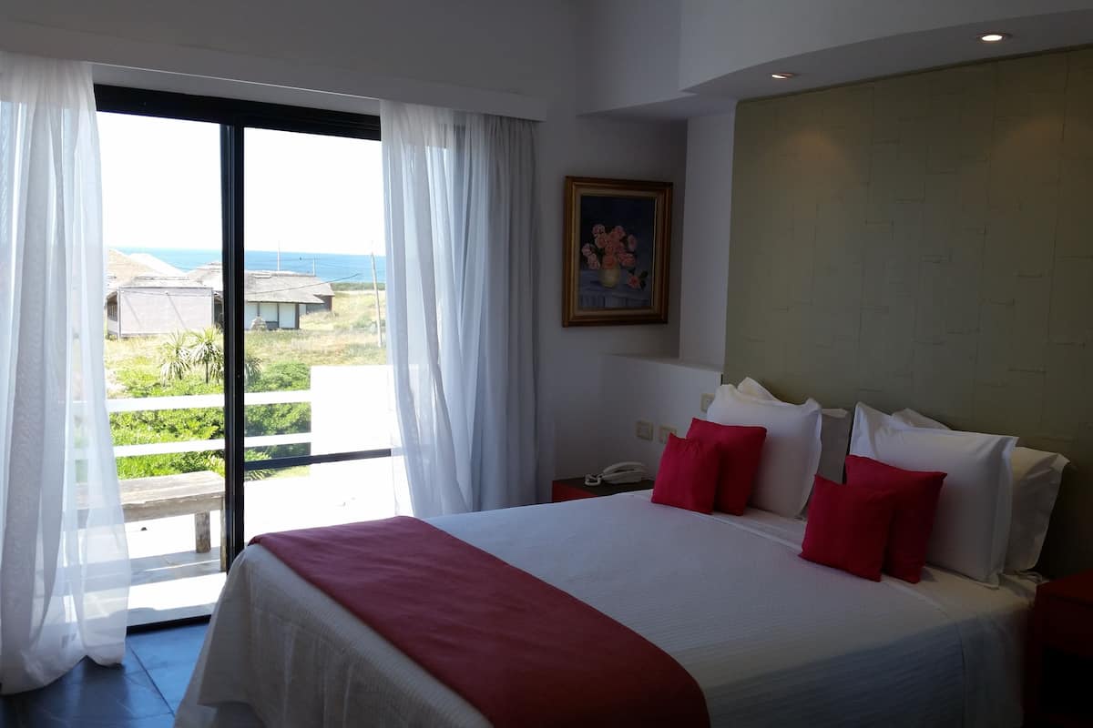 A hotel room with a large bed, white bedding, red pillows, a painting on the wall, and a view of the sea through a sliding door.