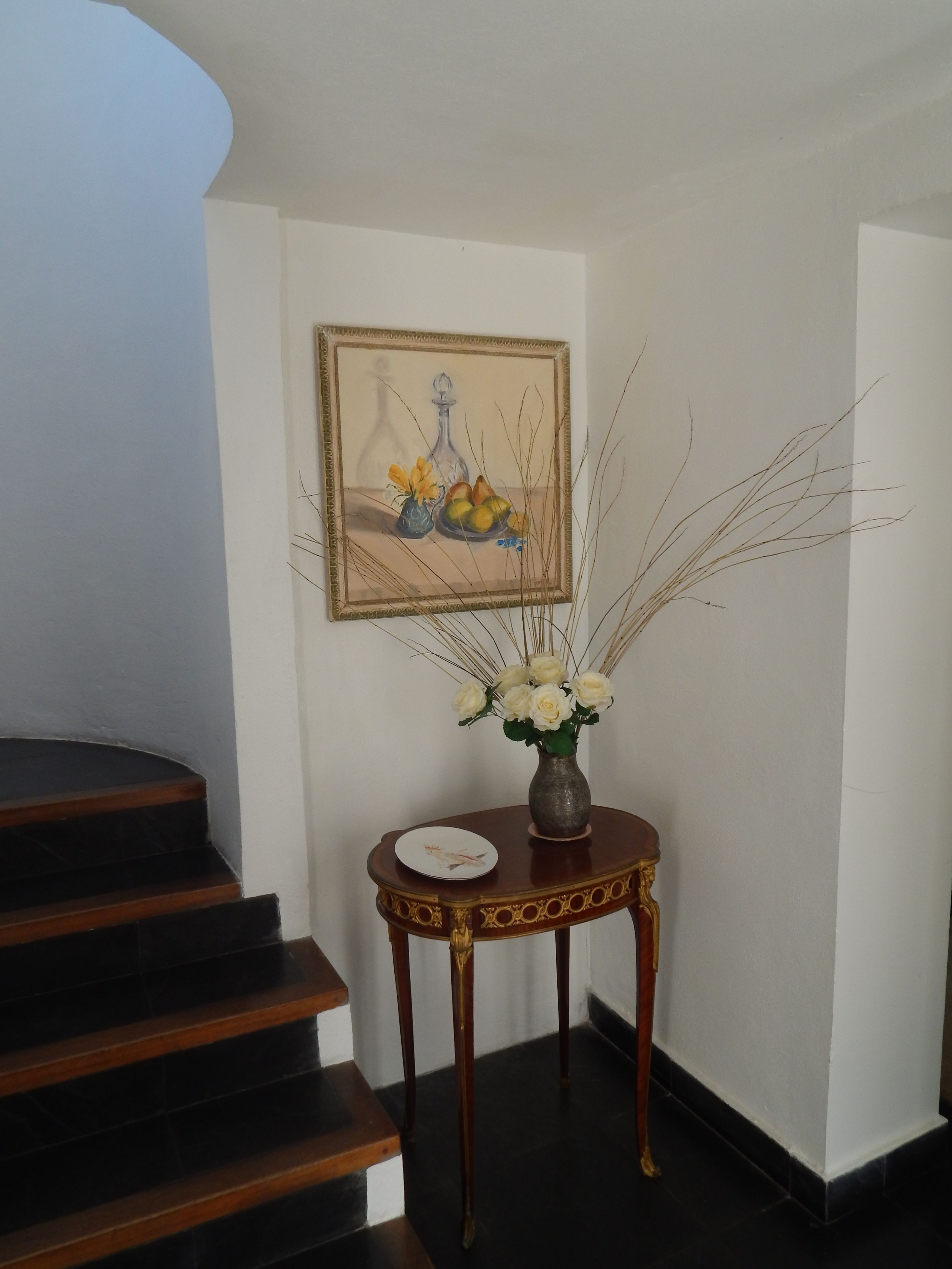 Una escalera con peldaños de madera oscura conduce a una plataforma. En la plataforma, hay una pequeña mesa redonda con un jarrón de flores blancas y un cuadro enmarcado con motivos de frutas y flores.