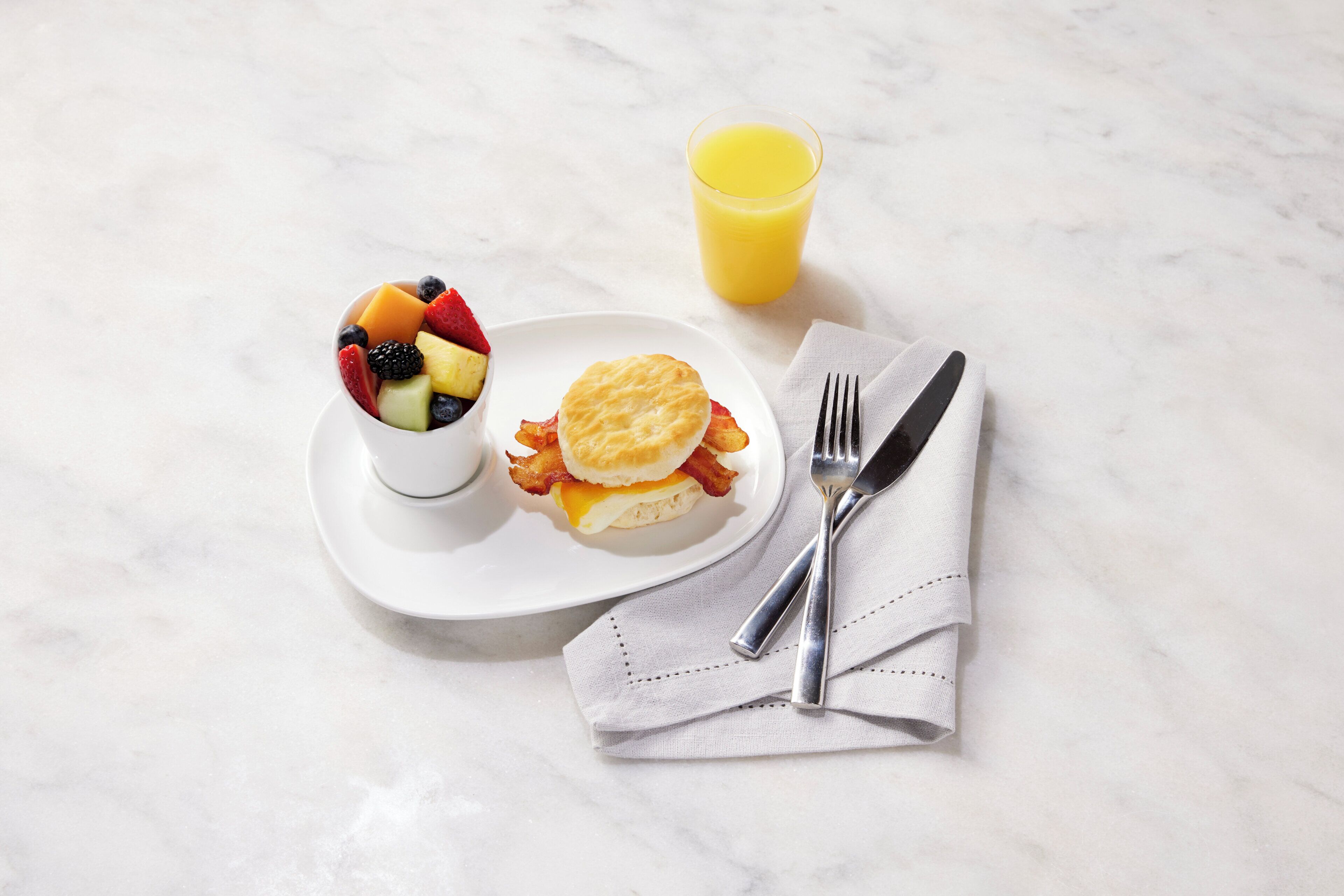 Un desayuno con un bol de fruta, un sándwich de galleta y un vaso de zumo de naranja sobre una superficie de mármol.