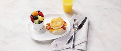 Un desayuno con un bol de fruta, un sándwich de galleta y un vaso de zumo de naranja sobre una superficie de mármol.