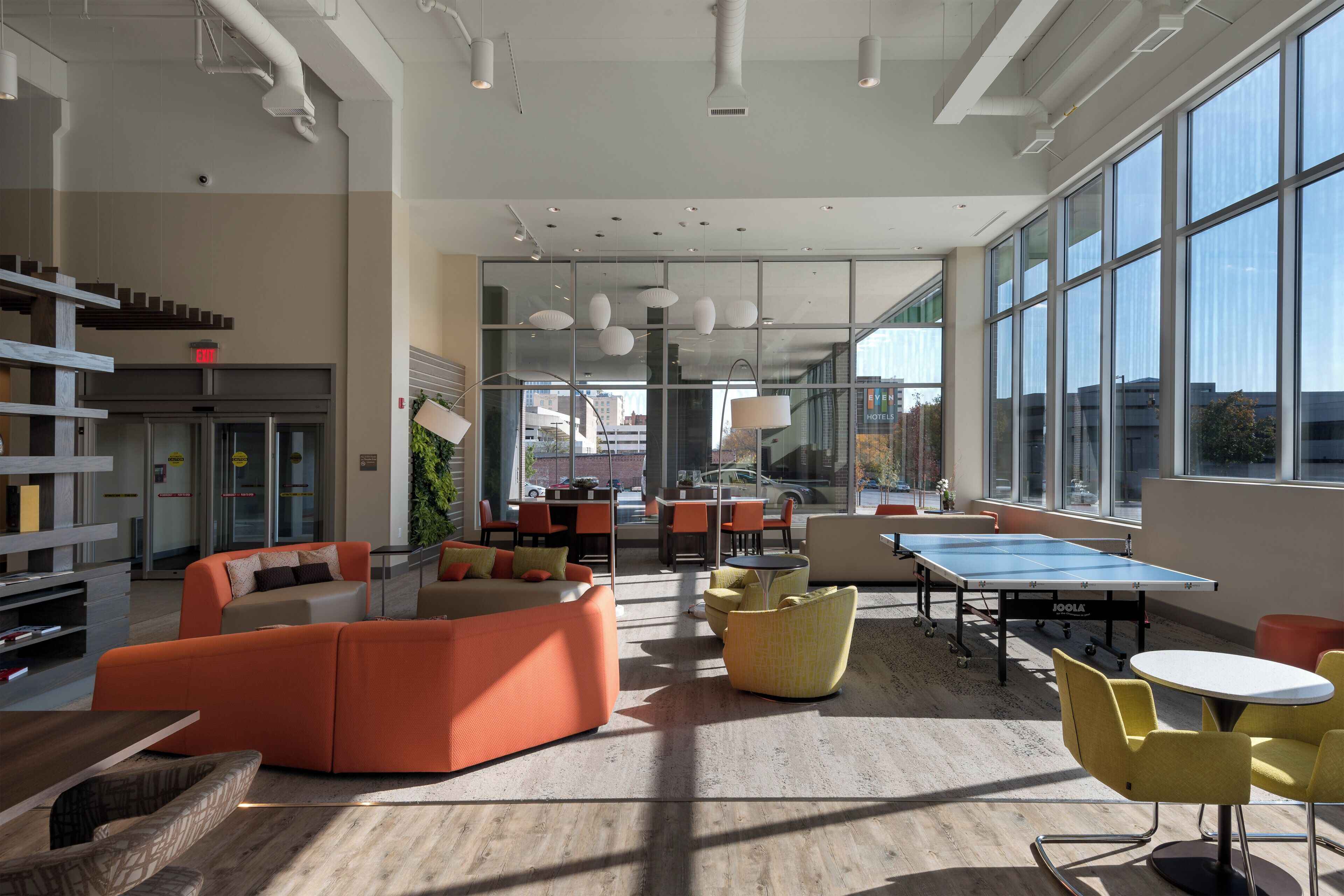 A modern hotel lobby with a ping pong table, orange and yellow seating, and large windows offering city views.