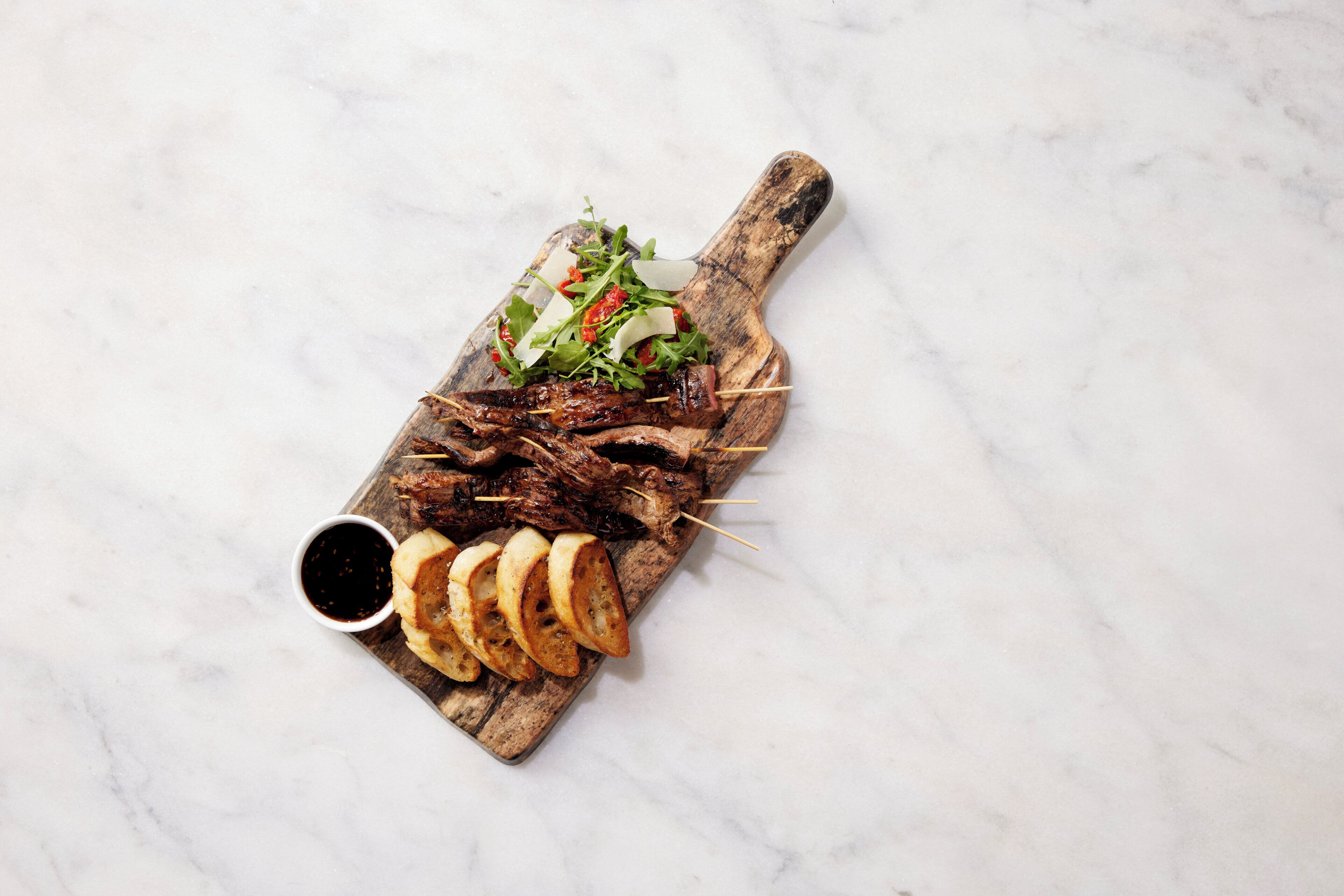 Une planche de service en bois, sur laquelle sont disposés des morceaux de viande en brochettes, des légumes verts et des tranches de pain, le tout posé sur une surface en marbre.