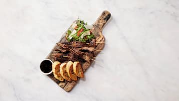 Une planche de service en bois, sur laquelle sont disposés des morceaux de viande en brochettes, des légumes verts et des tranches de pain, le tout posé sur une surface en marbre.