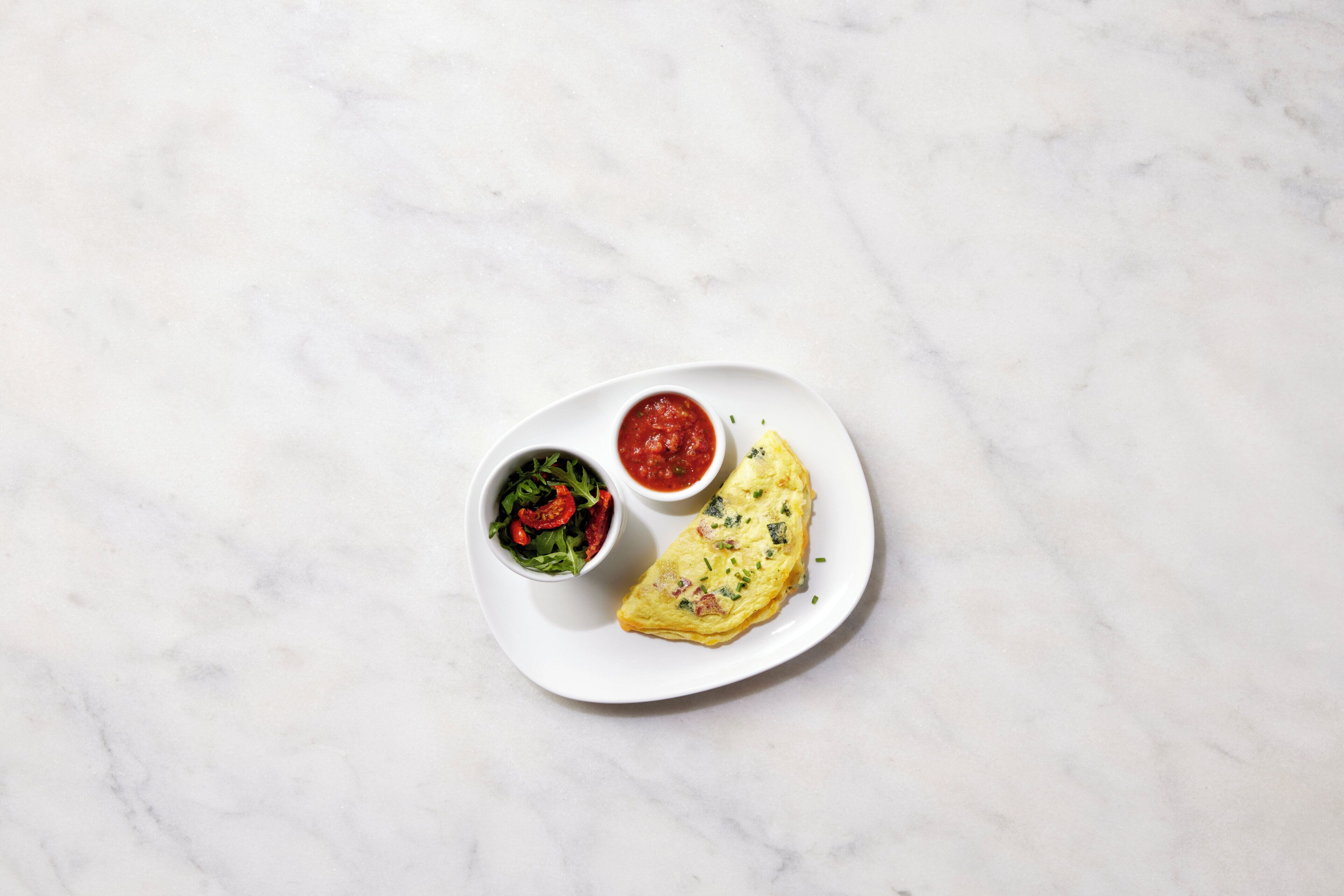 Une assiette blanche contenant une omelette, un bol de salade et un bol de sauce, le tout posé sur une surface en marbre.