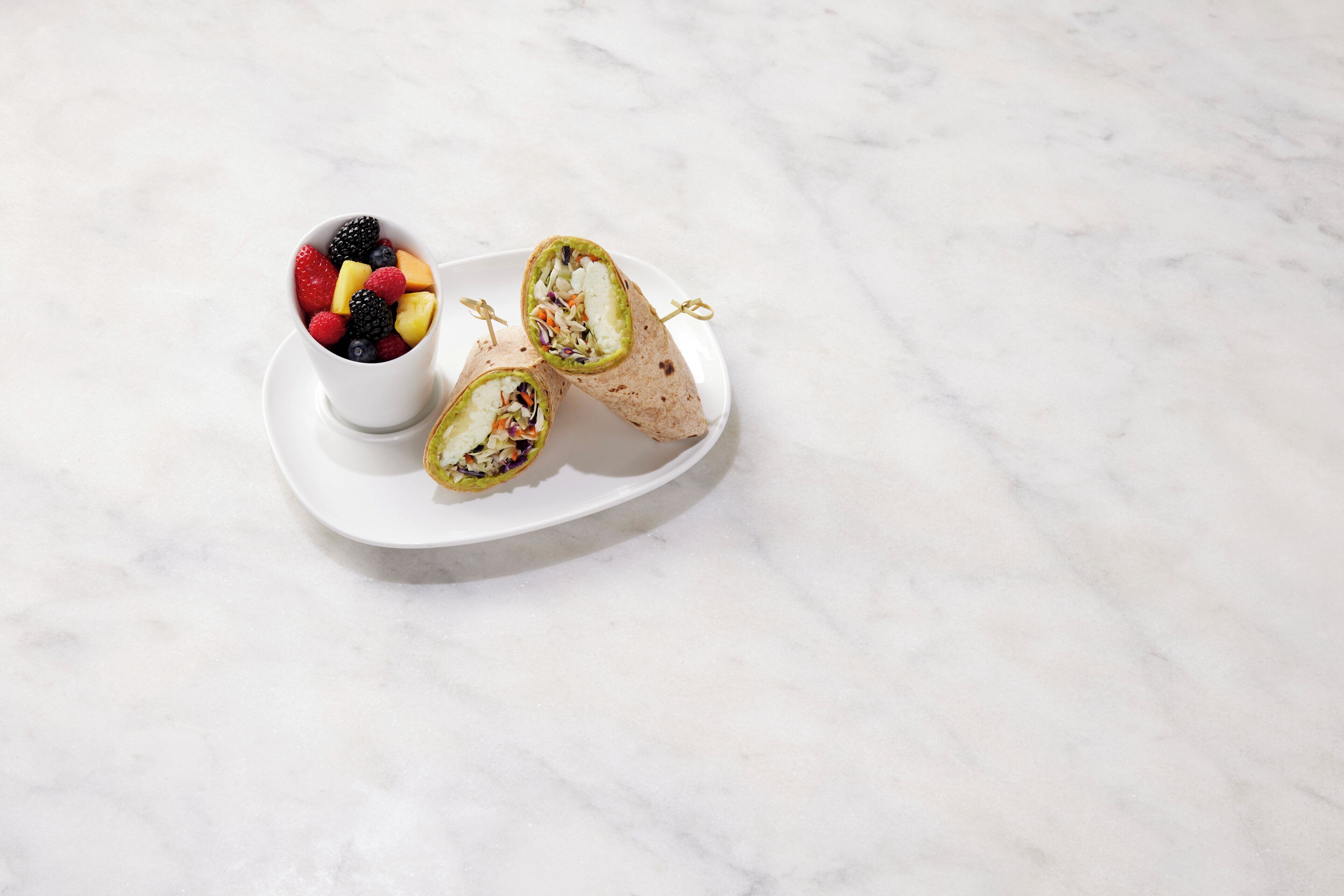 Een wit bord met een wrap en een bekertje fruit.