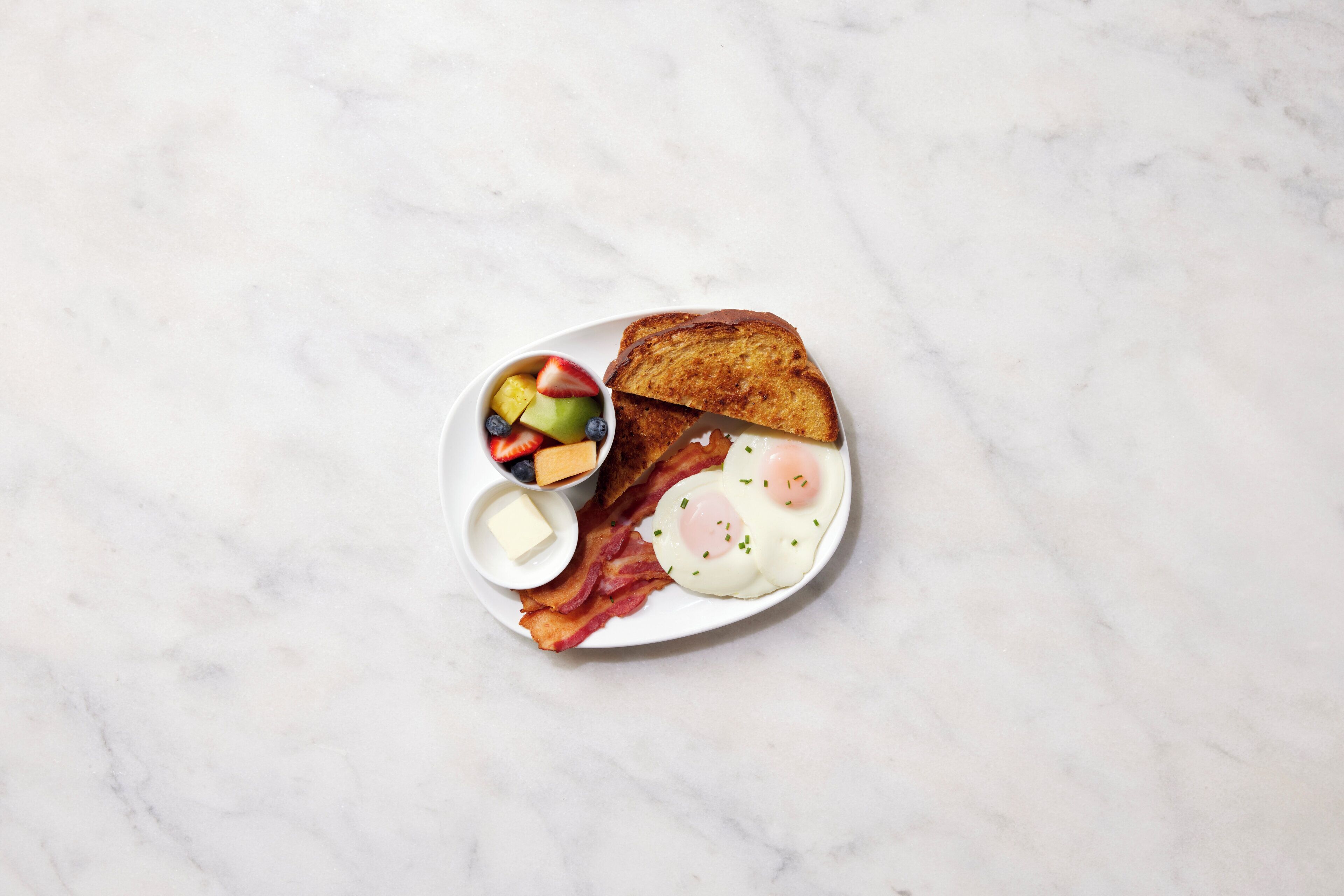 A breakfast plate with toast, eggs, bacon, and a fruit bowl.