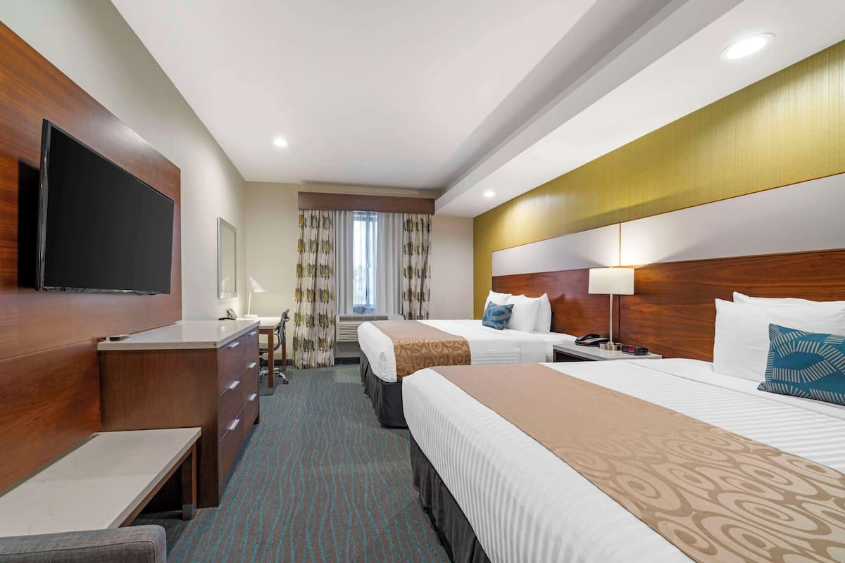 A hotel room with a large flat-screen TV, a desk with a chair, two beds with headboards, a bedside table with a lamp, and a window with curtains.