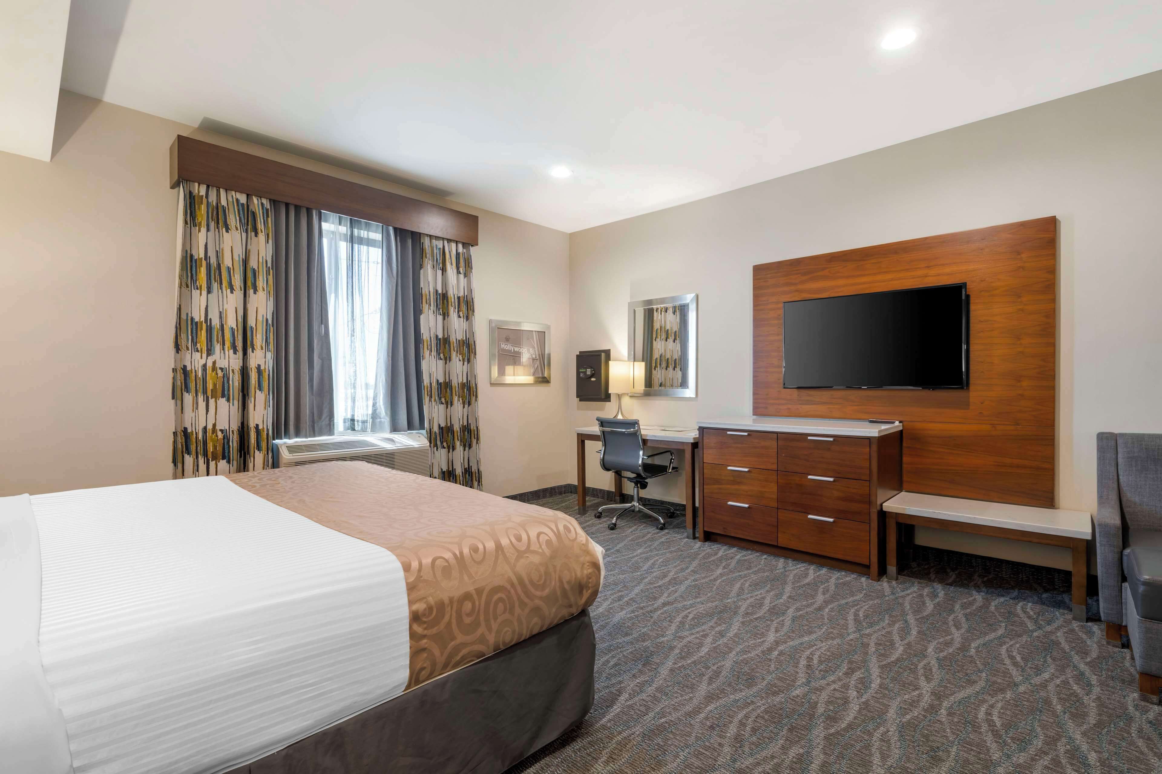 A hotel room with a large bed, a desk with a chair, a flat-screen TV mounted on the wall, and a bench.
