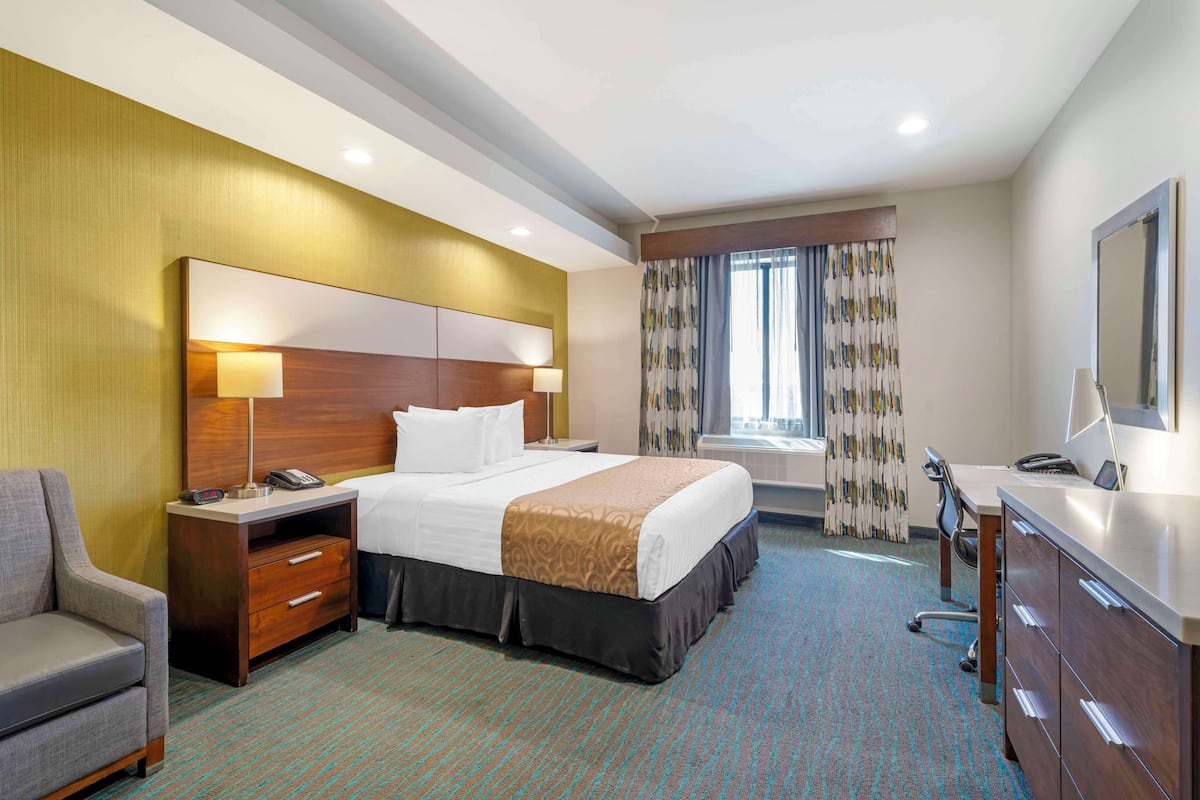 A hotel room with a large bed, a desk with a computer, a chair, a nightstand with a lamp, and a window with curtains.