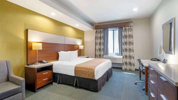 A hotel room with a large bed, a desk with a computer, a chair, a nightstand with a lamp, and a window with curtains.