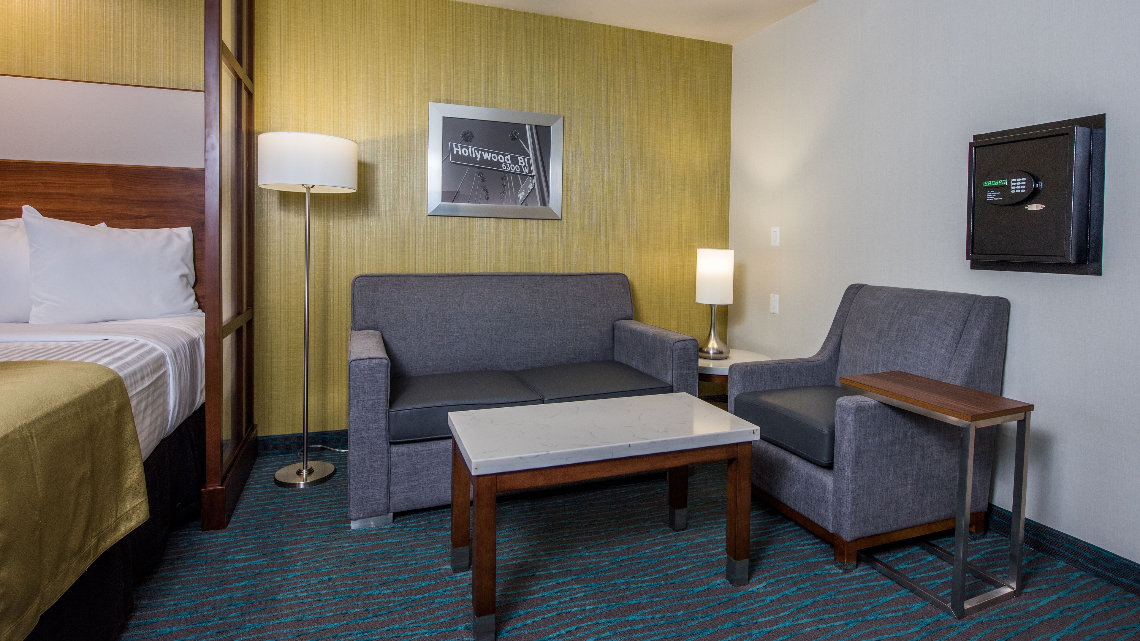 A hotel room with a bed, a gray sofa, a small table, and a chair. There is a framed picture on the wall and a lamp on the side table.
