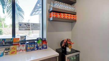 A hotel reception area with a counter displaying various products like Advil, Motrin, and cereal boxes. There are shelves above the counter holding water bottles and a small refrigerator with a Coca-Cola bottle inside.