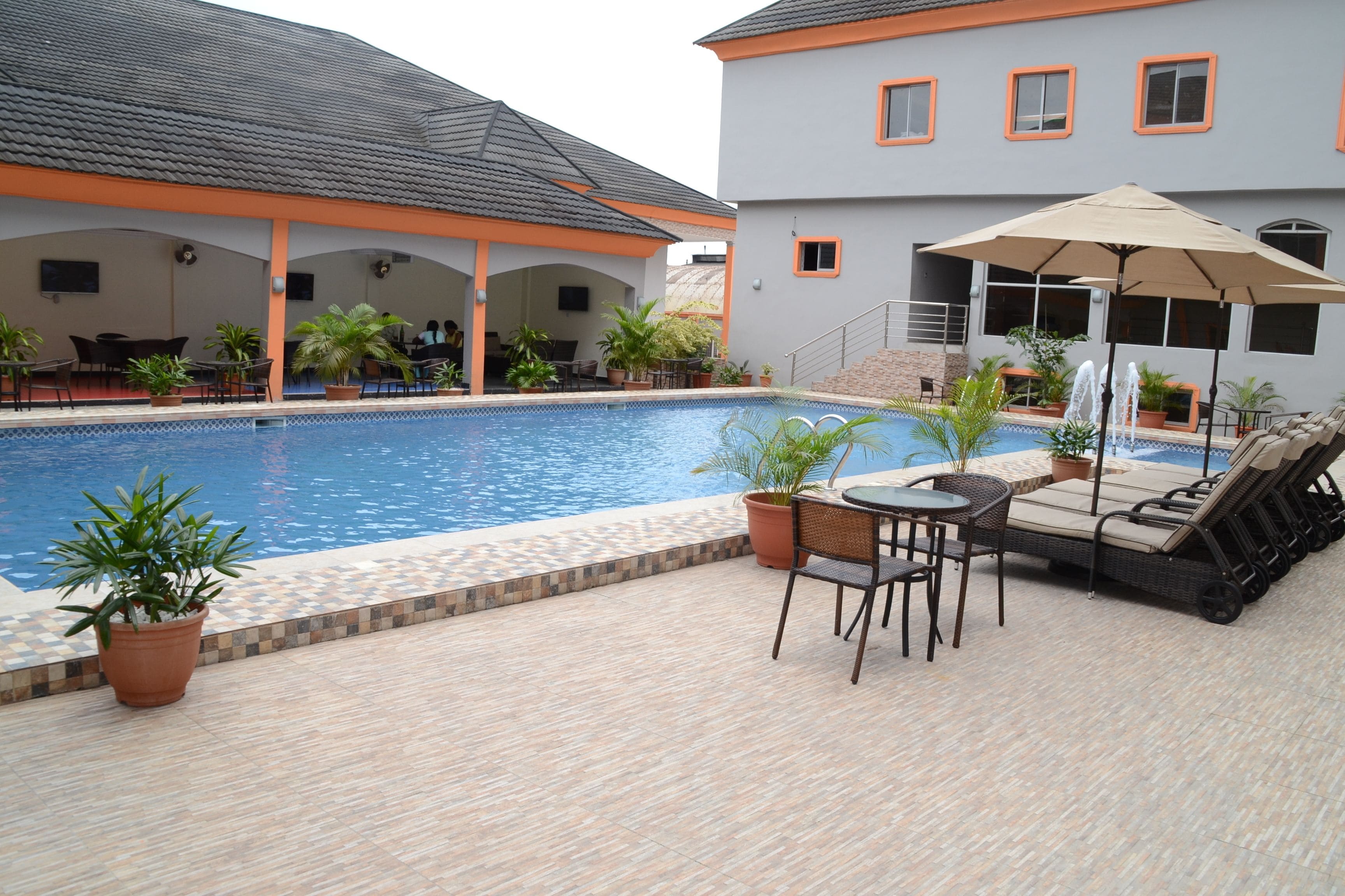 A pool area with lounge chairs and a table, surrounded by potted plants and a covered seating area.