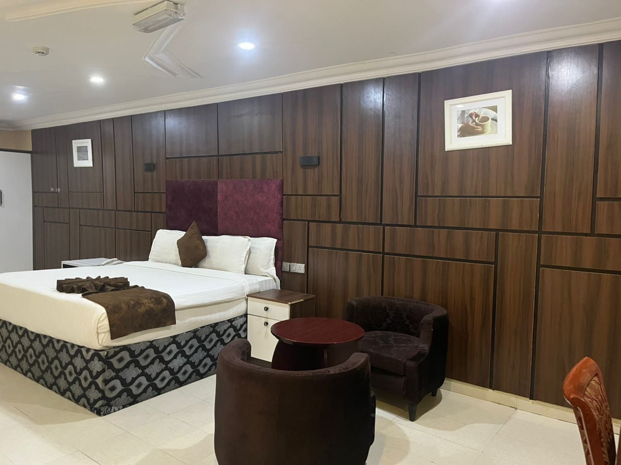 A hotel room with a bed, bedside table, two chairs, a small round table, and a wooden panel wall.
