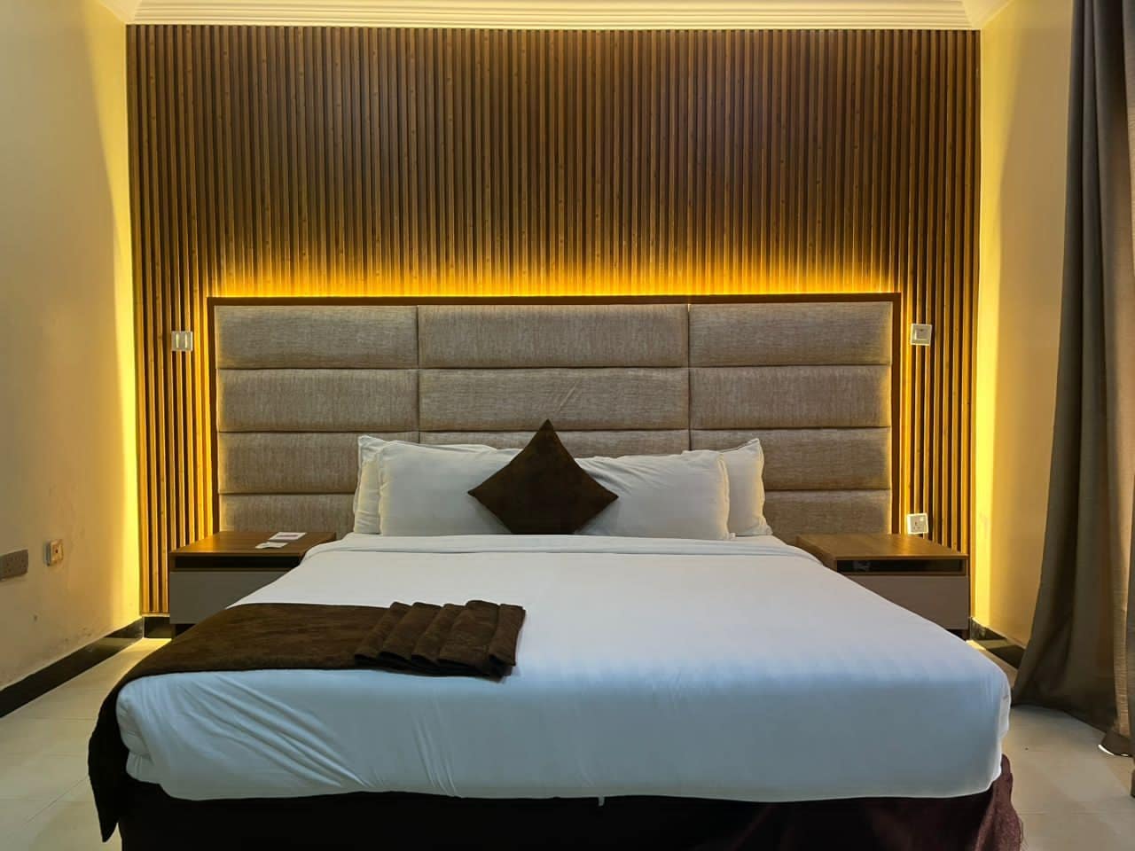 Una habitación de hotel con una cama grande, un cabecero con líneas verticales y una mesita de noche.
