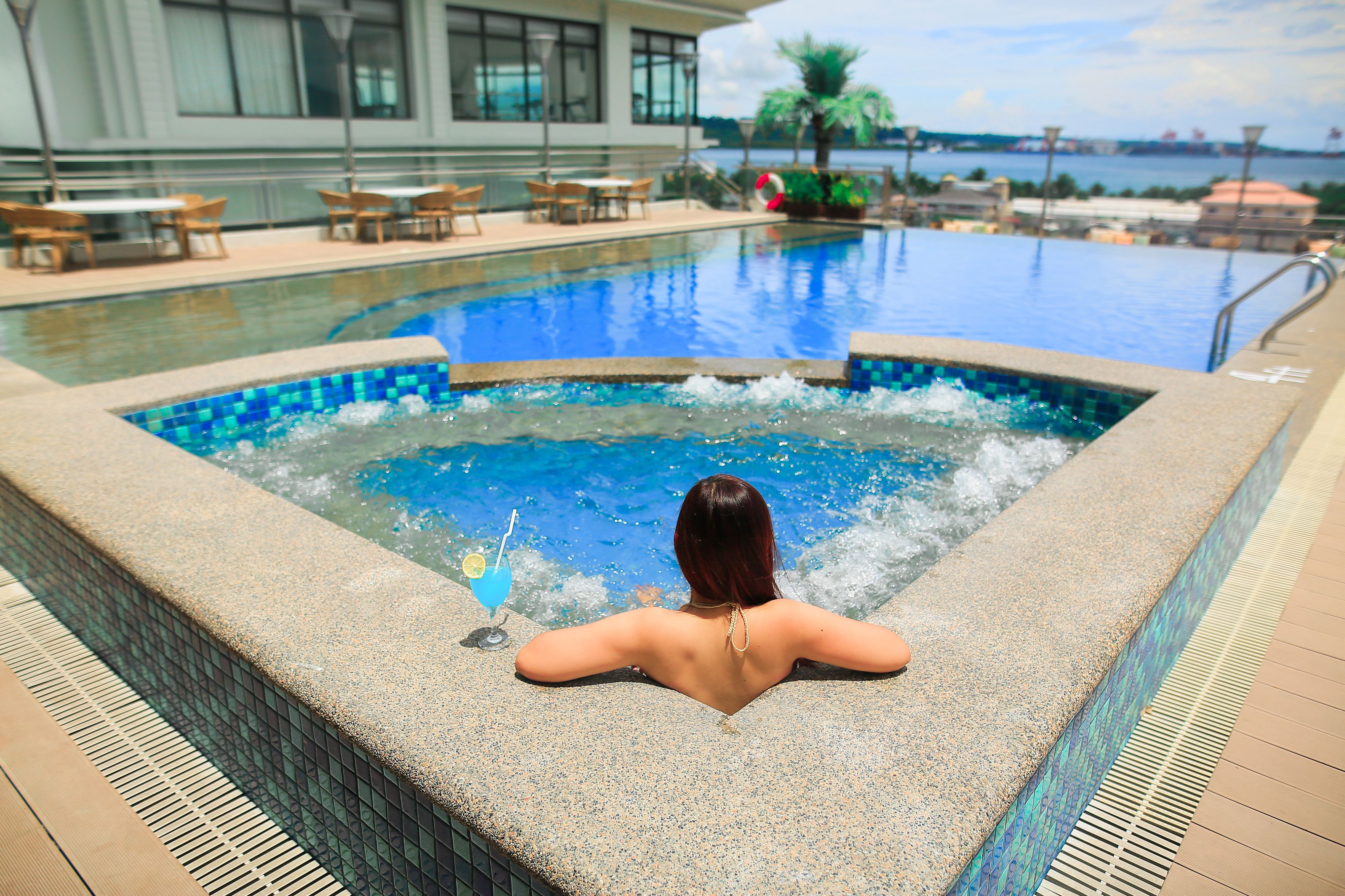 A woman relaxing in a hot tub at a poolside area with a view of the sea and a building in the background.