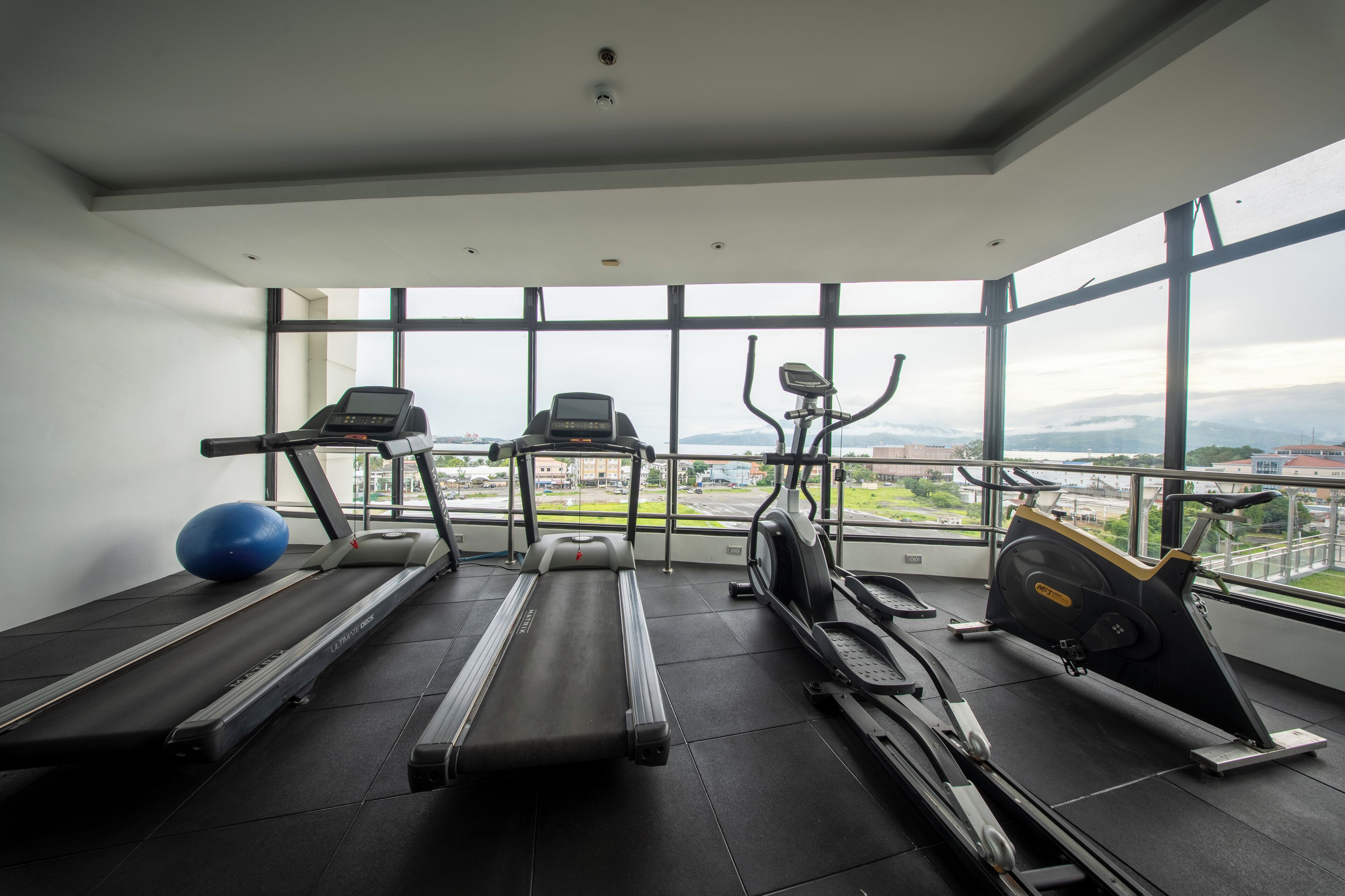 A gym with treadmills, an elliptical, and a stationary bike, large windows with a city view.