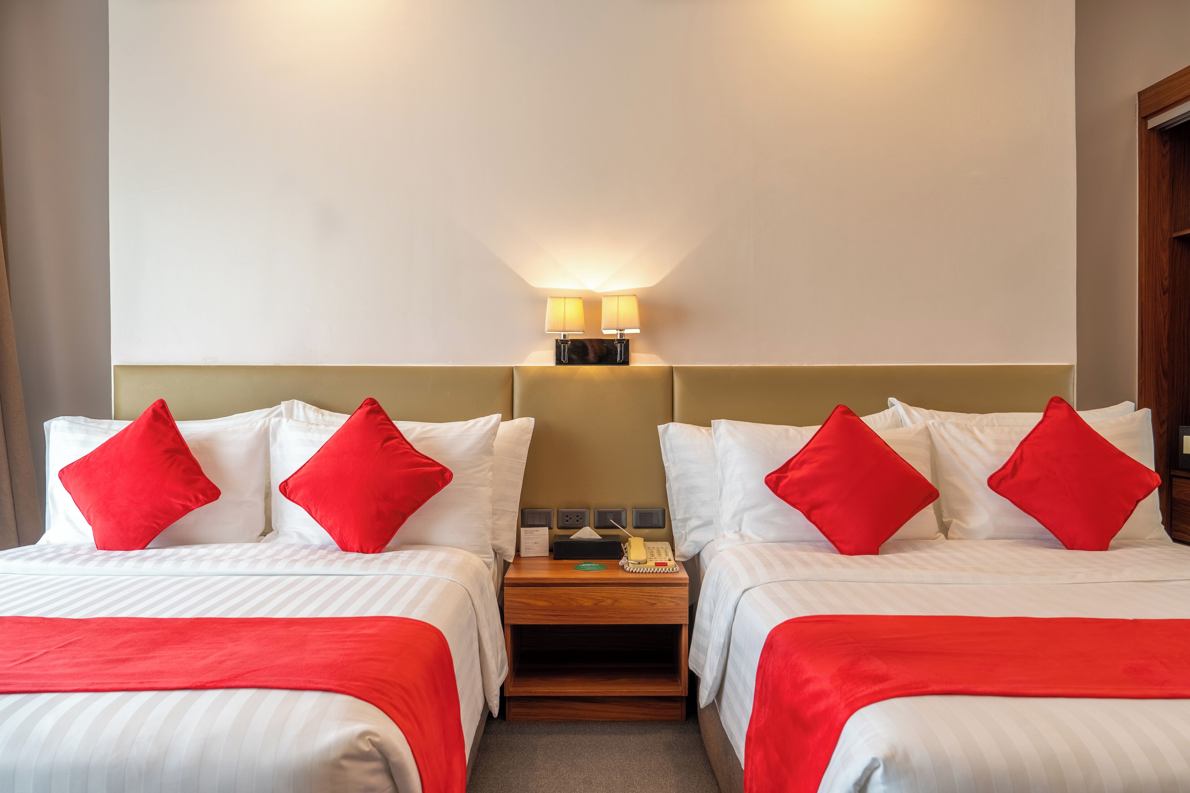 A hotel room with two beds, each with white bedding and red pillows. There is a nightstand between the beds with a lamp and a telephone.