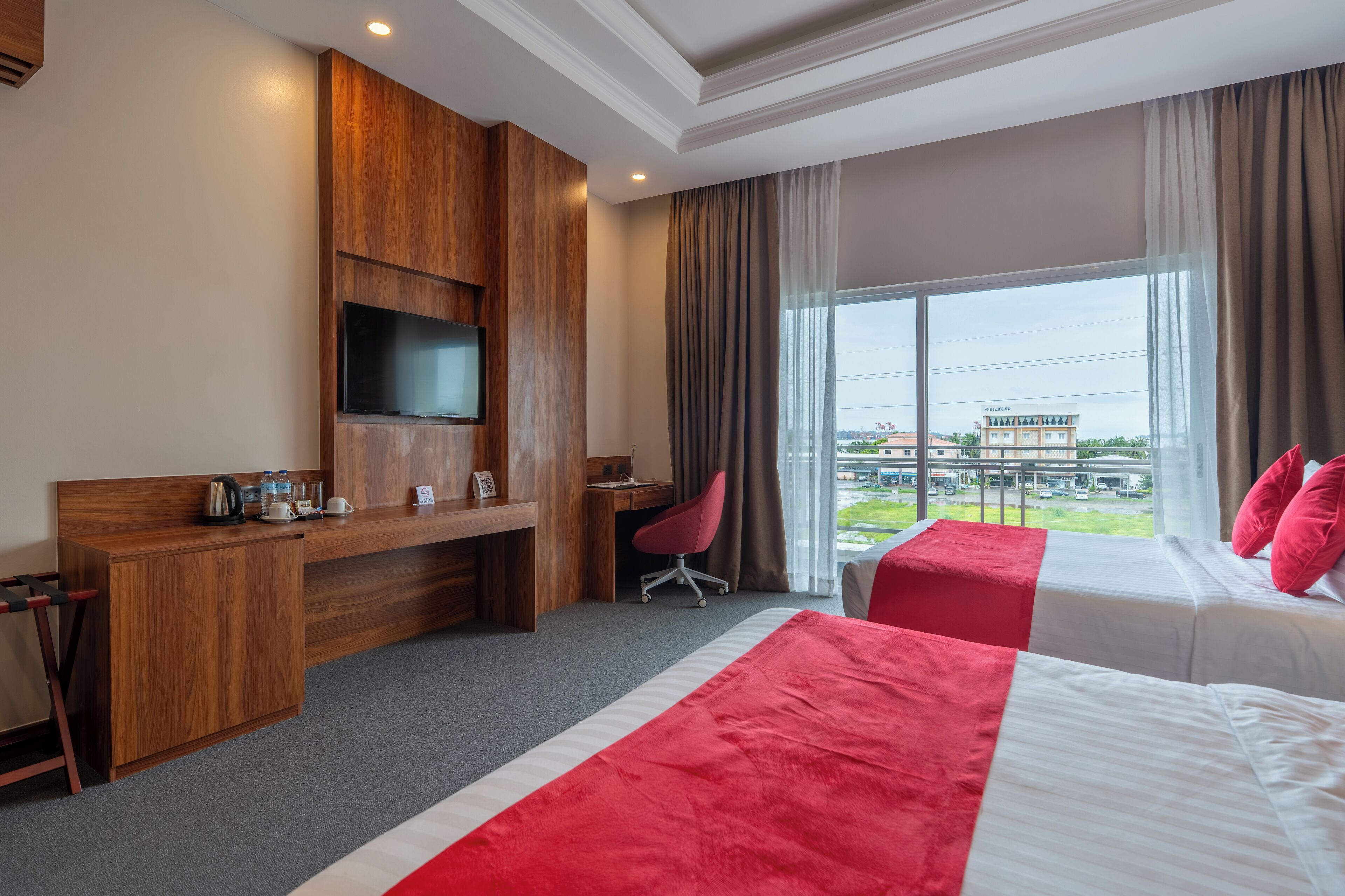 A hotel room with a wooden desk, a TV, a bed with red and white bedding, and a large window with a view of buildings outside.