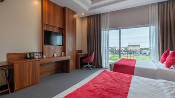 A hotel room with a wooden desk, a TV, a bed with red and white bedding, and a large window with a view of buildings outside.