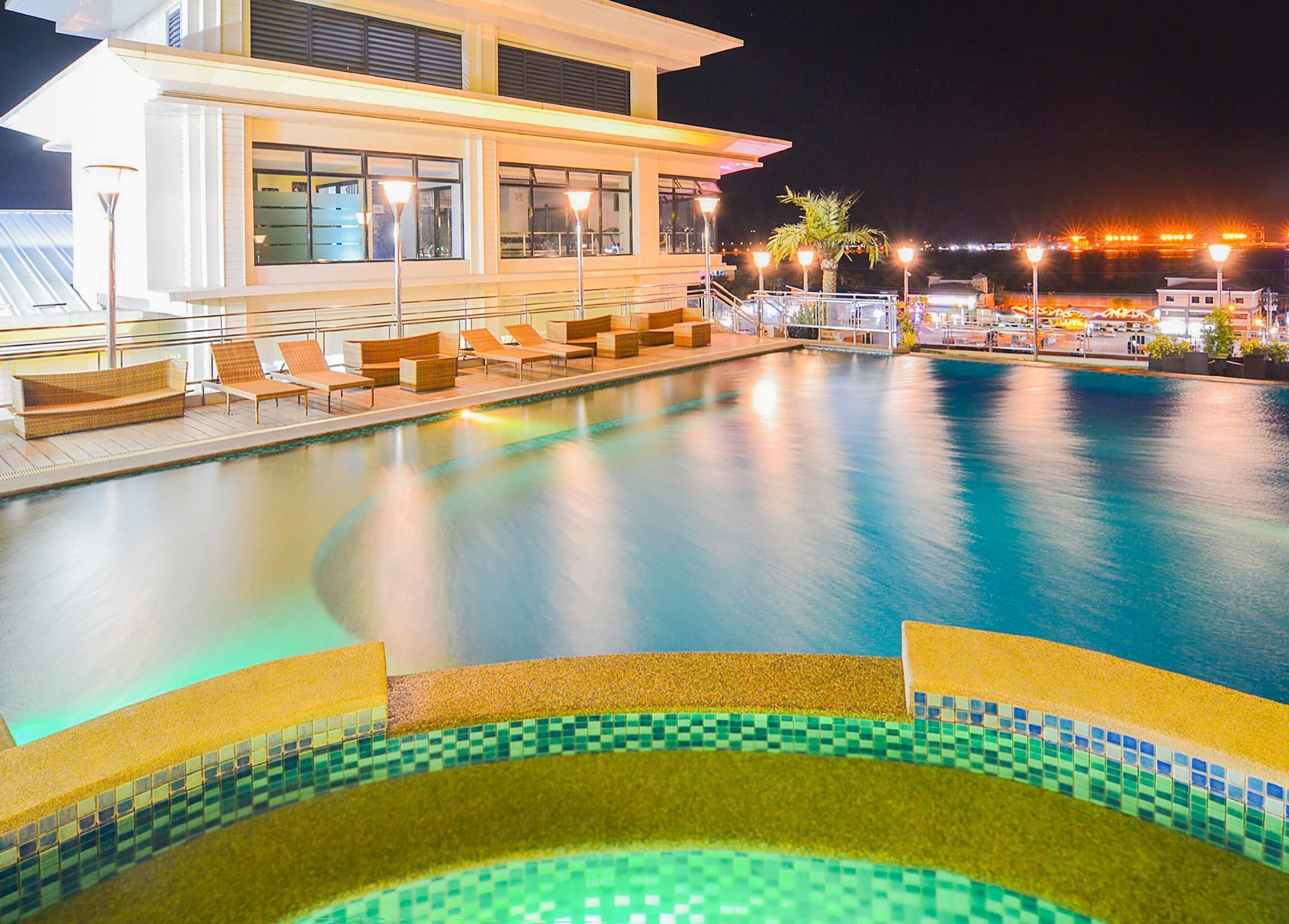 A swimming pool with lounge chairs and a tiled edge, surrounded by a building with large windows and outdoor lighting.