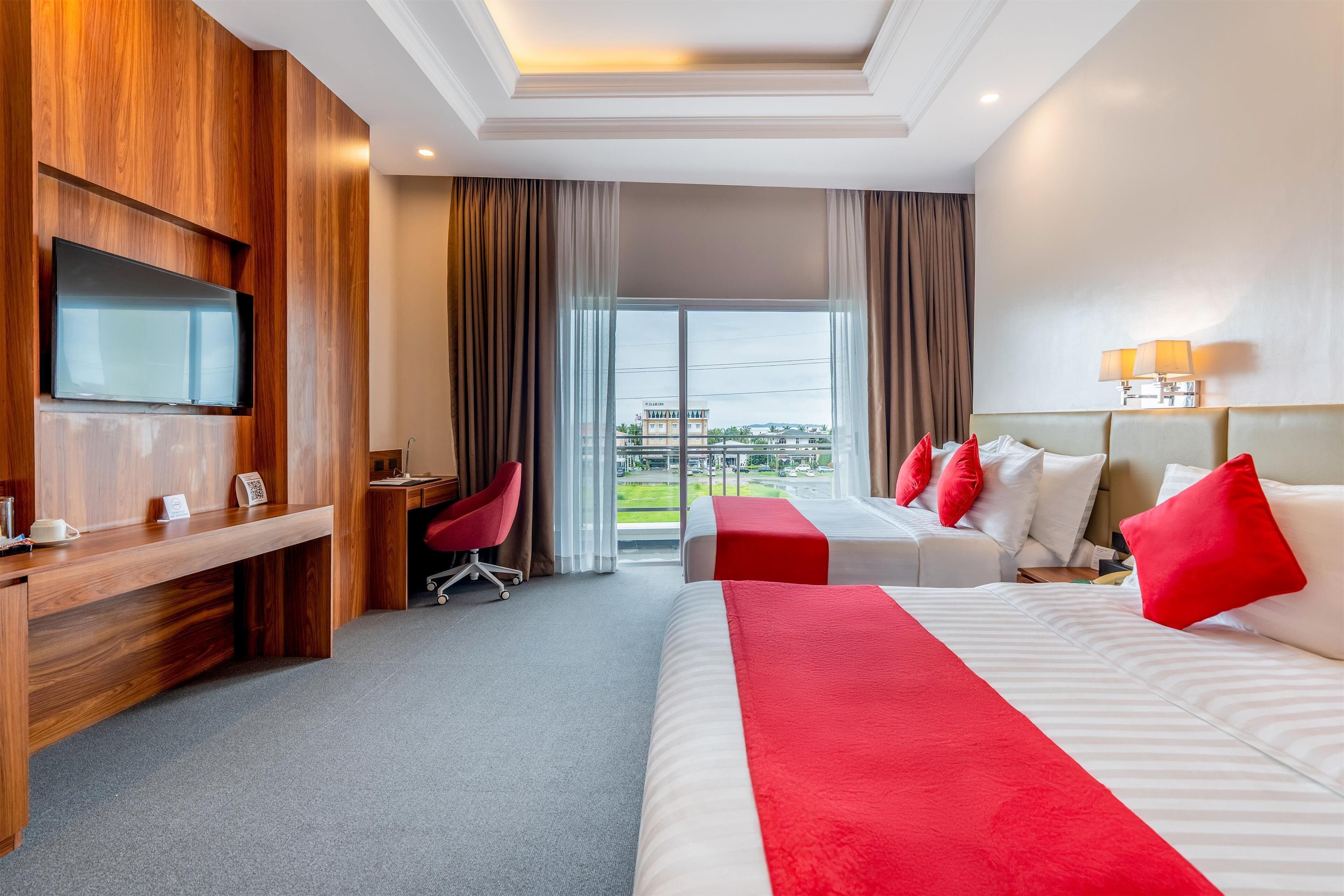 A hotel room with a wooden desk, a TV, two beds with red pillows, a chair, and a view of the outdoors.
