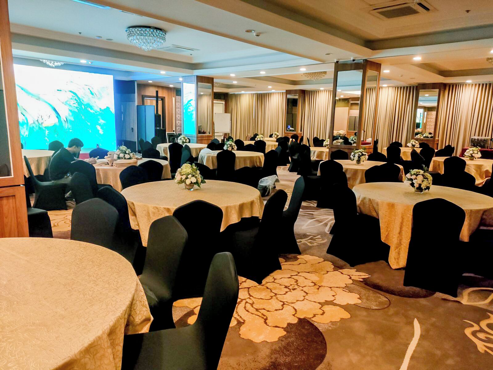 Sala de conferência com mesas redondas, cadeiras pretas e centros de mesa florais.