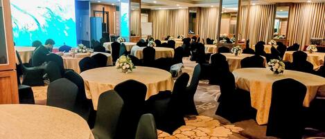 Sala de conferência com mesas redondas, cadeiras pretas e centros de mesa florais.