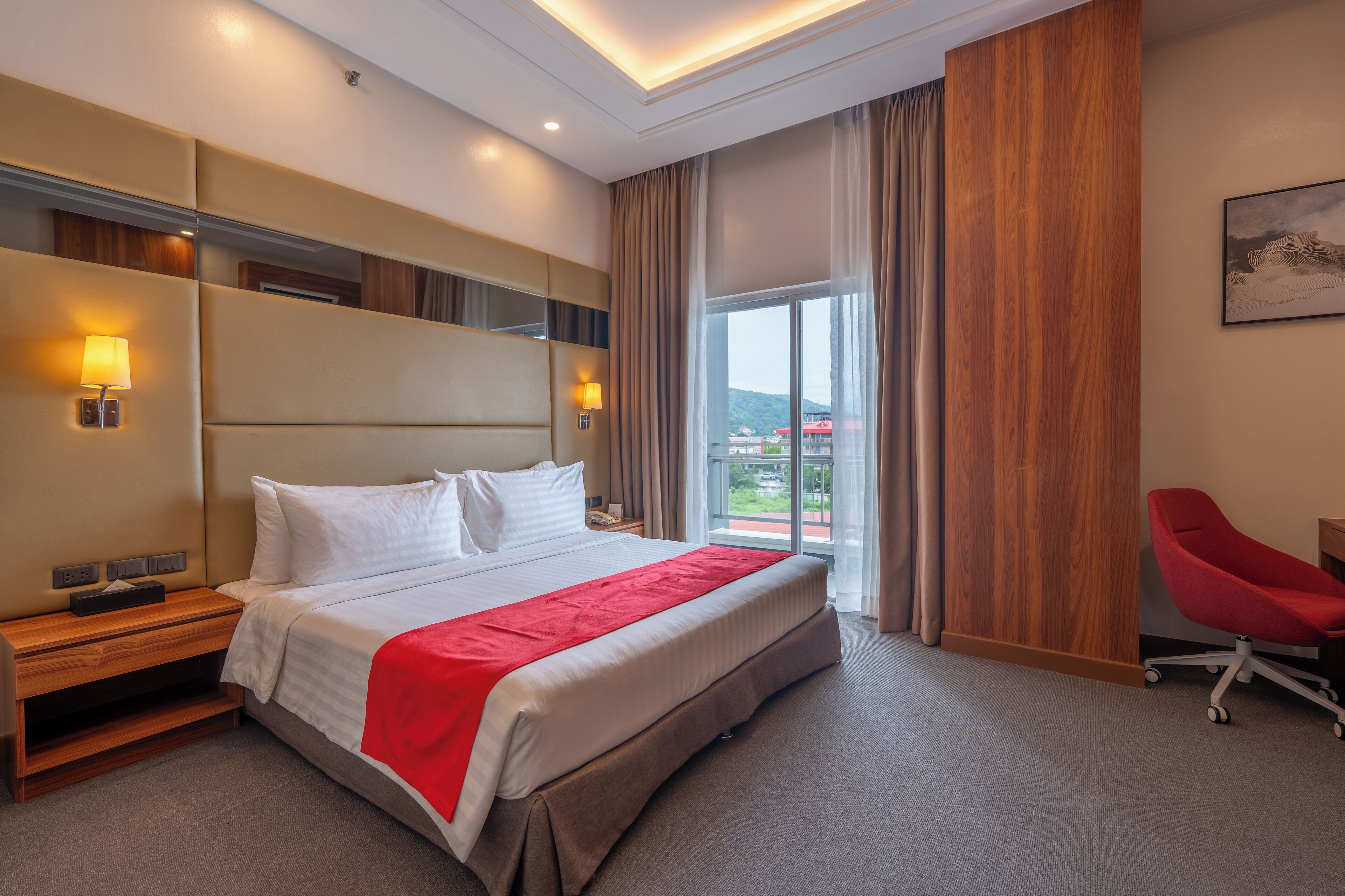A hotel room with a large bed, bedside tables, a red chair, and a view of the outdoors.