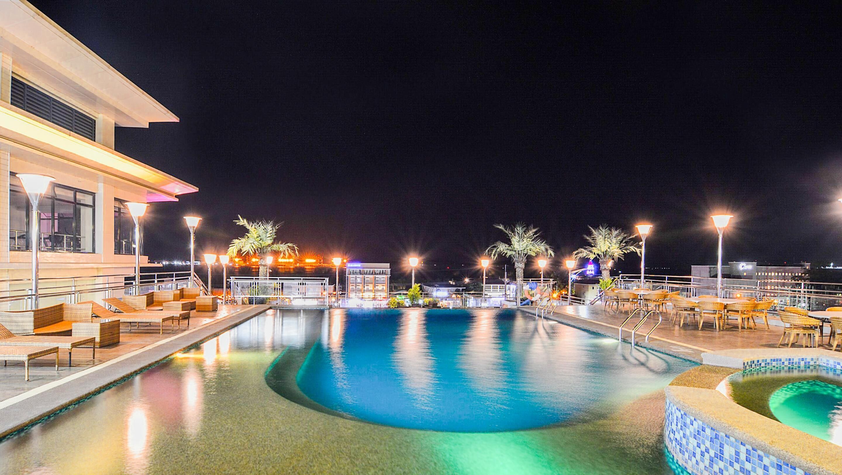 Ein Poolbereich bei Nacht mit Liegestühlen und Tischen, einem Pool und einem Gebäude mit beleuchteten Fenstern.