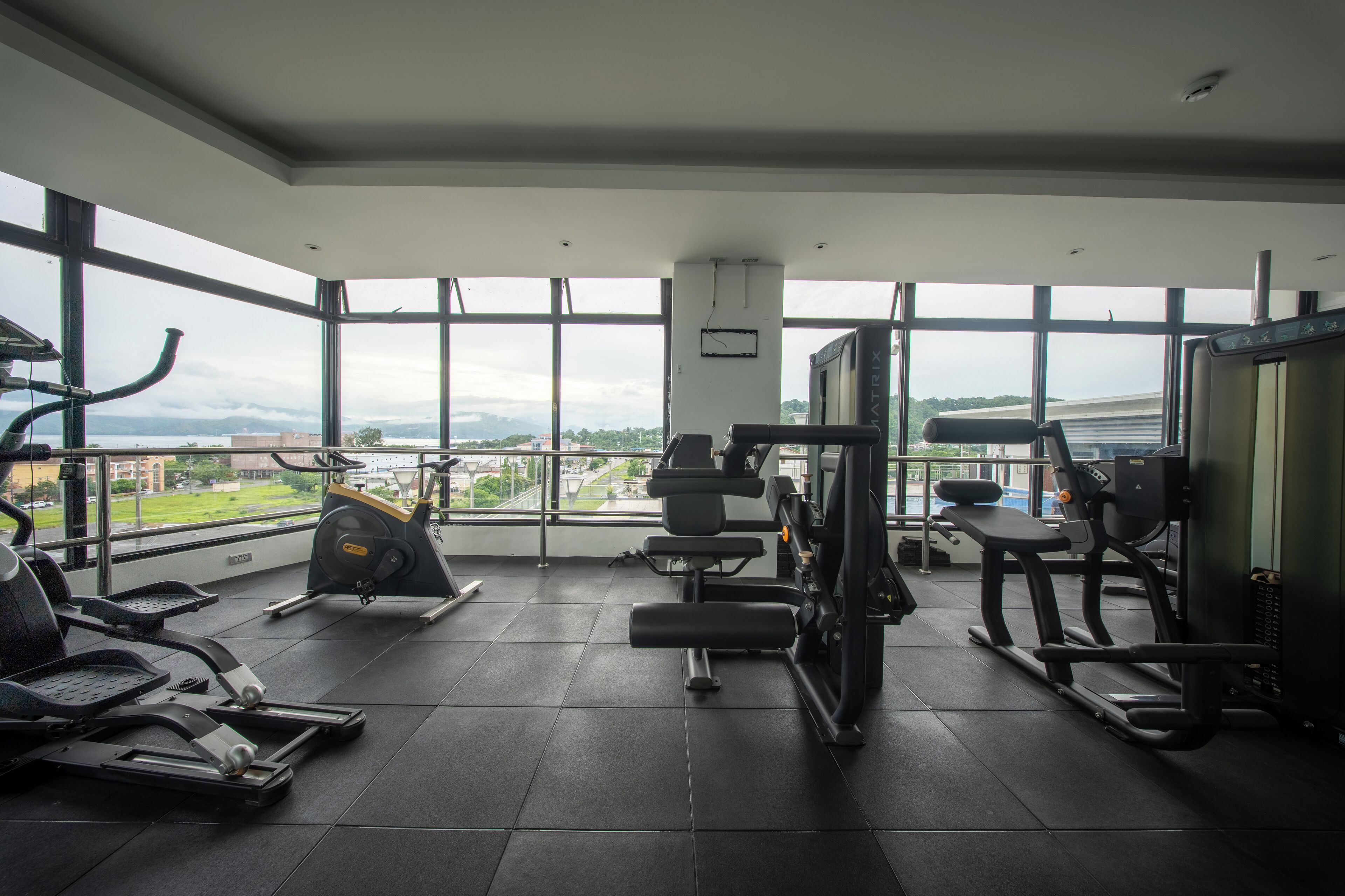 Un gimnasio con diferentes máquinas de ejercicio, ventanas amplias y vistas a un paisaje urbano.