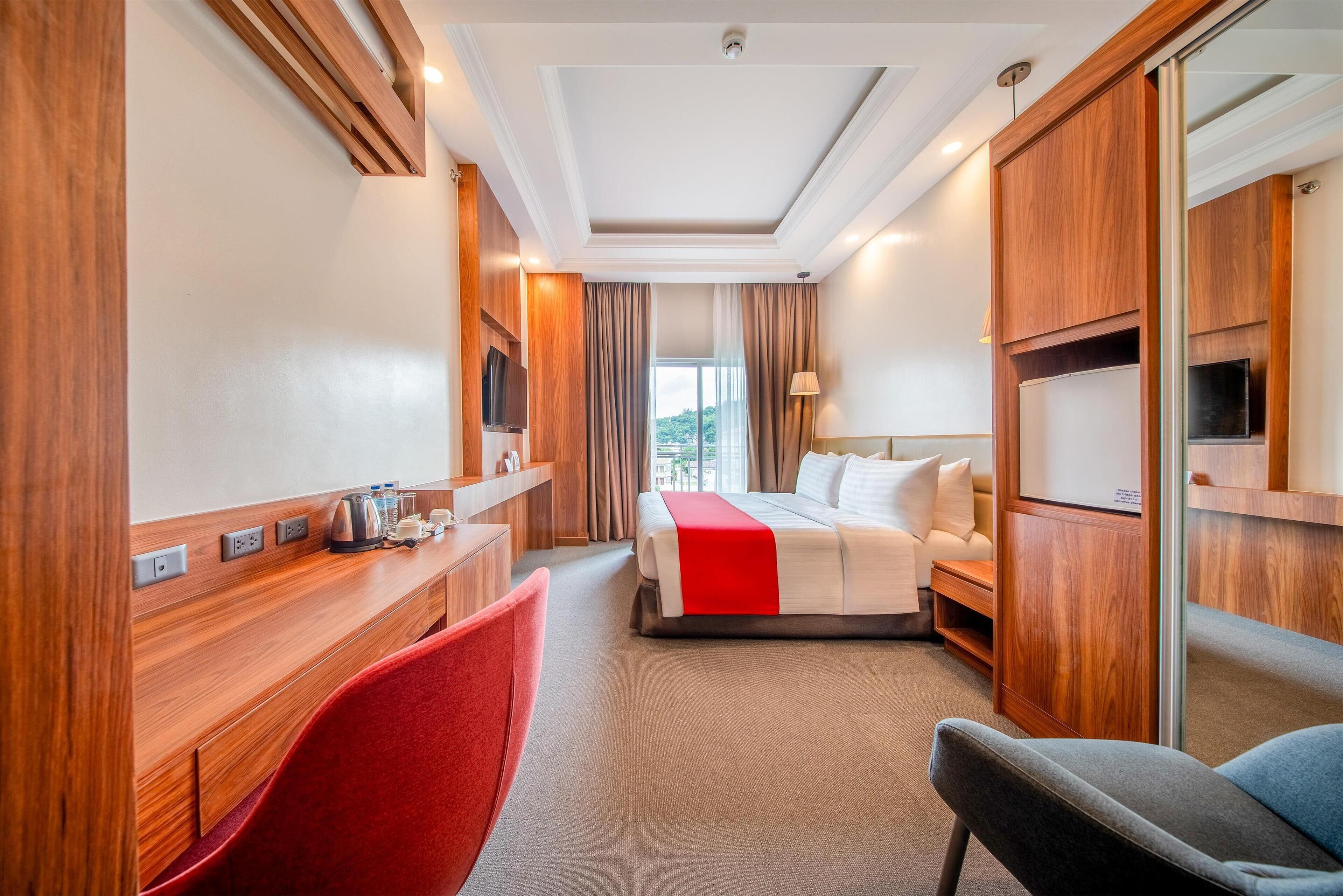A hotel room with a bed, a desk with a red chair, a TV, and a large mirror.