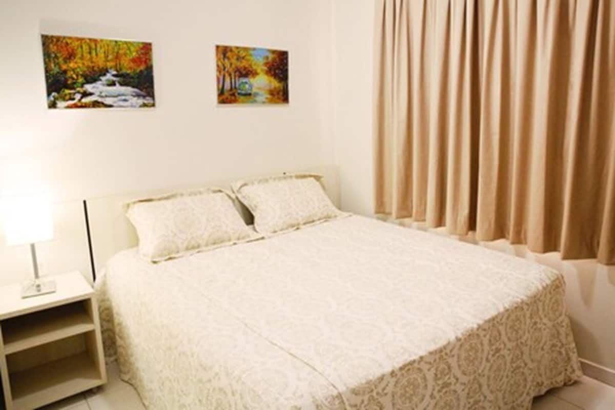 Um quarto com uma cama, duas pinturas na parede, uma mesa de cabeceira e cortinas bege.